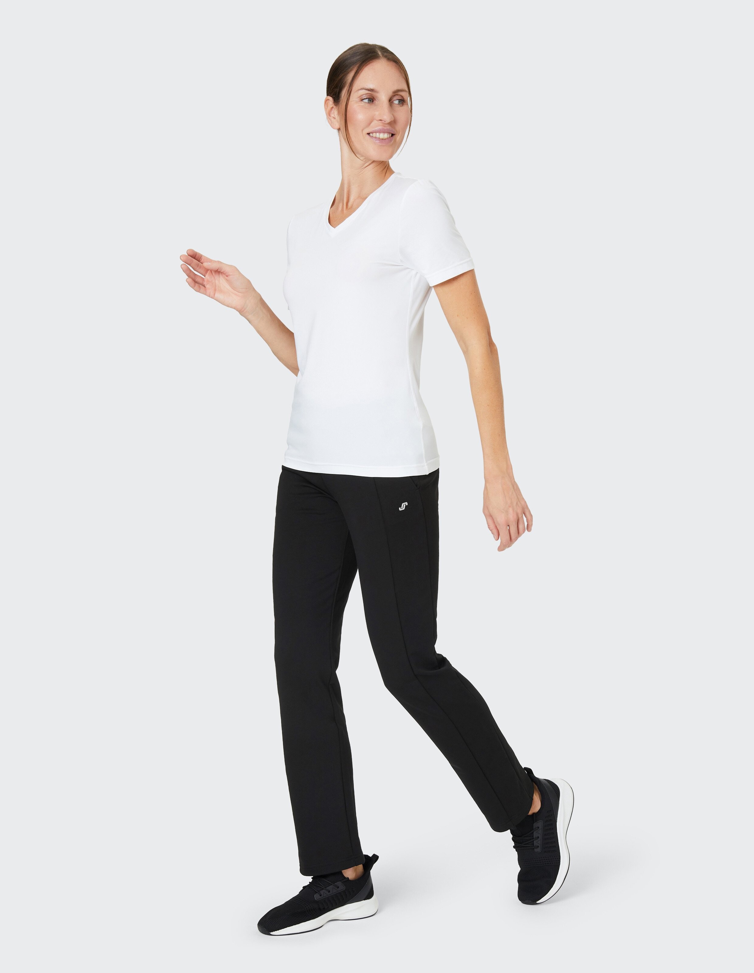 Frau in weißem T-Shirt und schwarzen Sporthosen läuft lächelnd, trägt schwarze Sneakers, Hintergrund ist neutral grau.