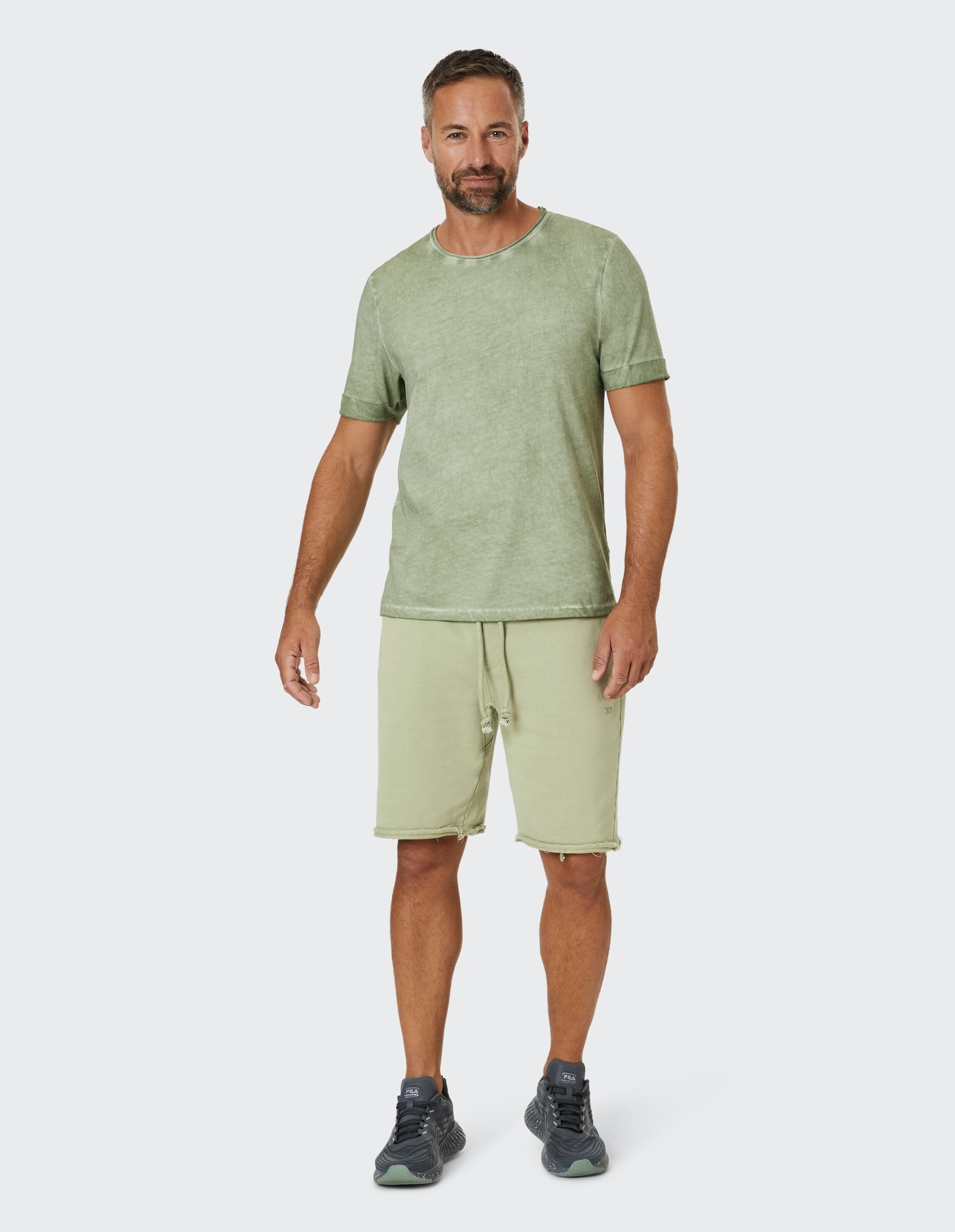 Ein Mann trägt ein pastellgrünes T-Shirt und passende Shorts, steht lächelnd und entspannt in grau-blauen Sportschuhen.