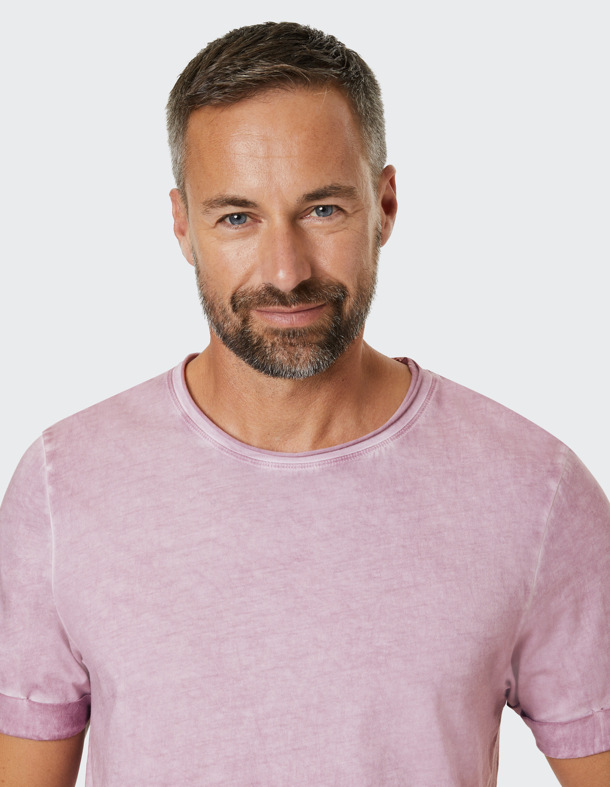 Mann mit kurzem grauem Haar und Bart trägt ein rosafarbenes T-Shirt und lächelt leicht vor einem hellen Hintergrund.