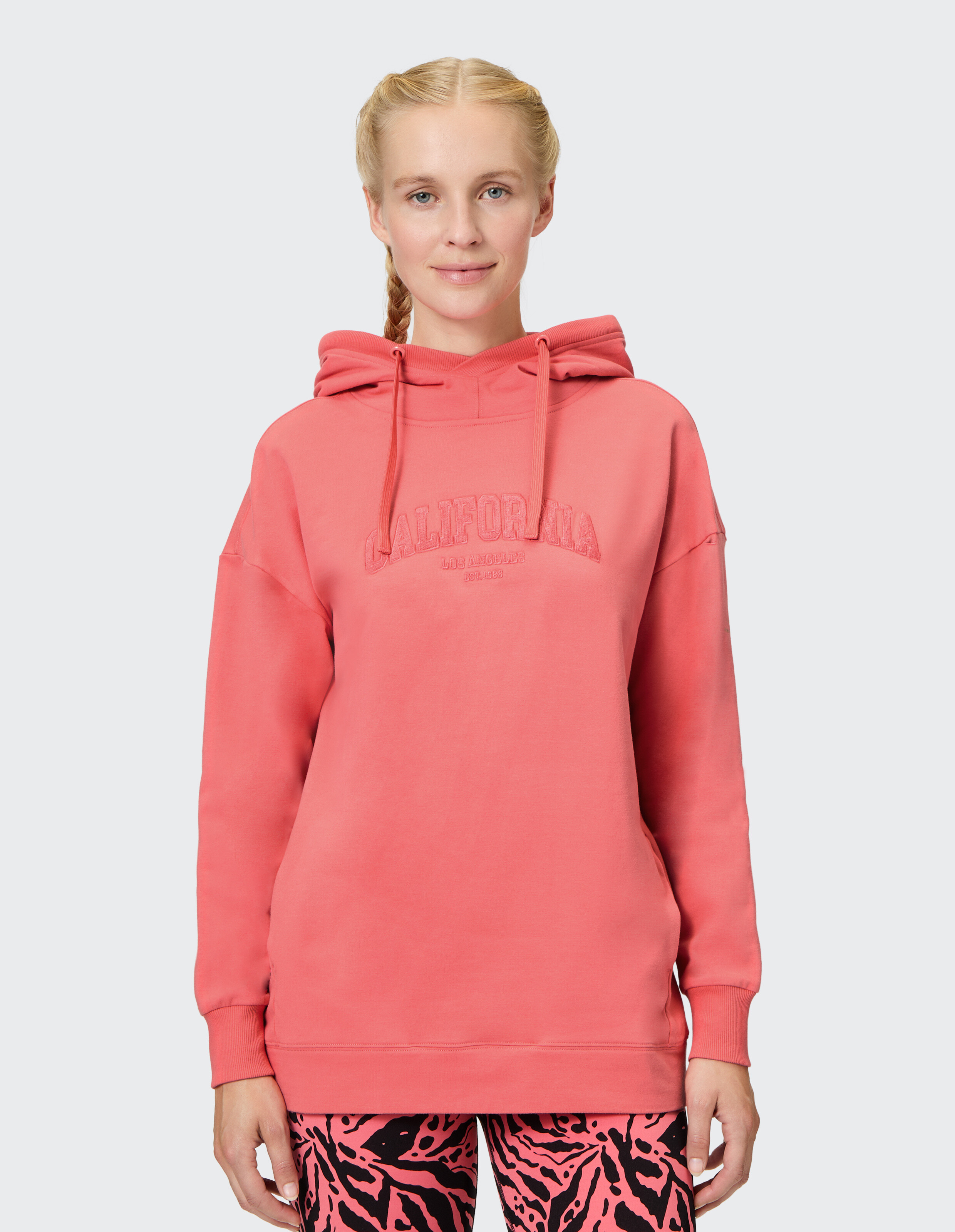 Frau trägt einen gemütlichen korallenroten Venice Beach Hoodie Muriel mit „California“-Schriftzug.