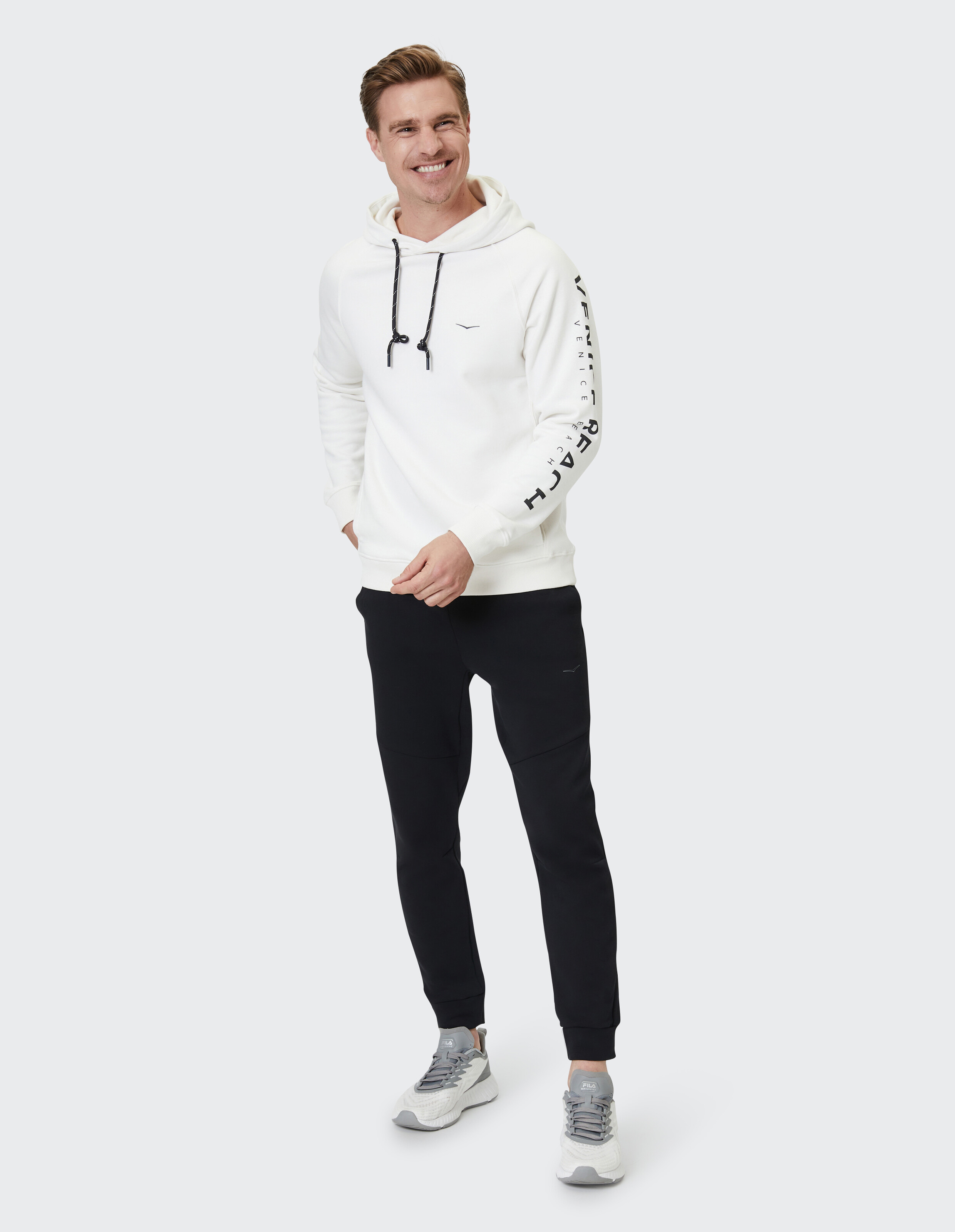 Mann trägt einen weißen ELBSAND-Hoodie mit Ärmelaufdruck und eine schwarze lässige Jogginghose.