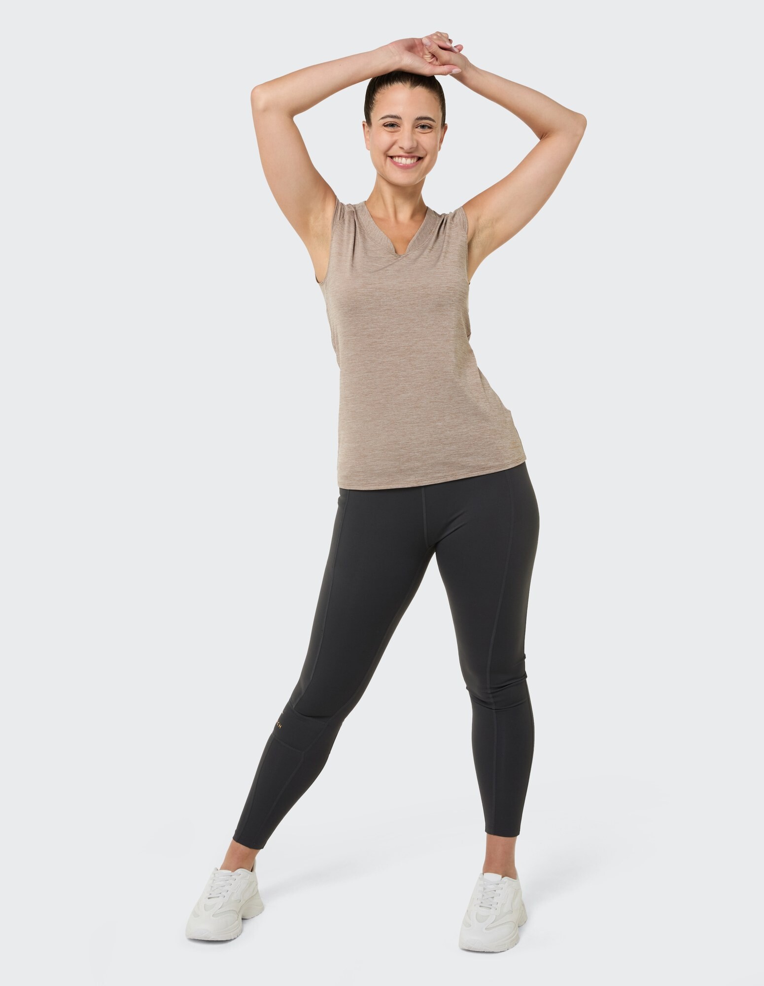 Lächelnde Frau in einem beigen ärmellosen Top und schwarzen Leggings, die Freizeitkleidung präsentiert.