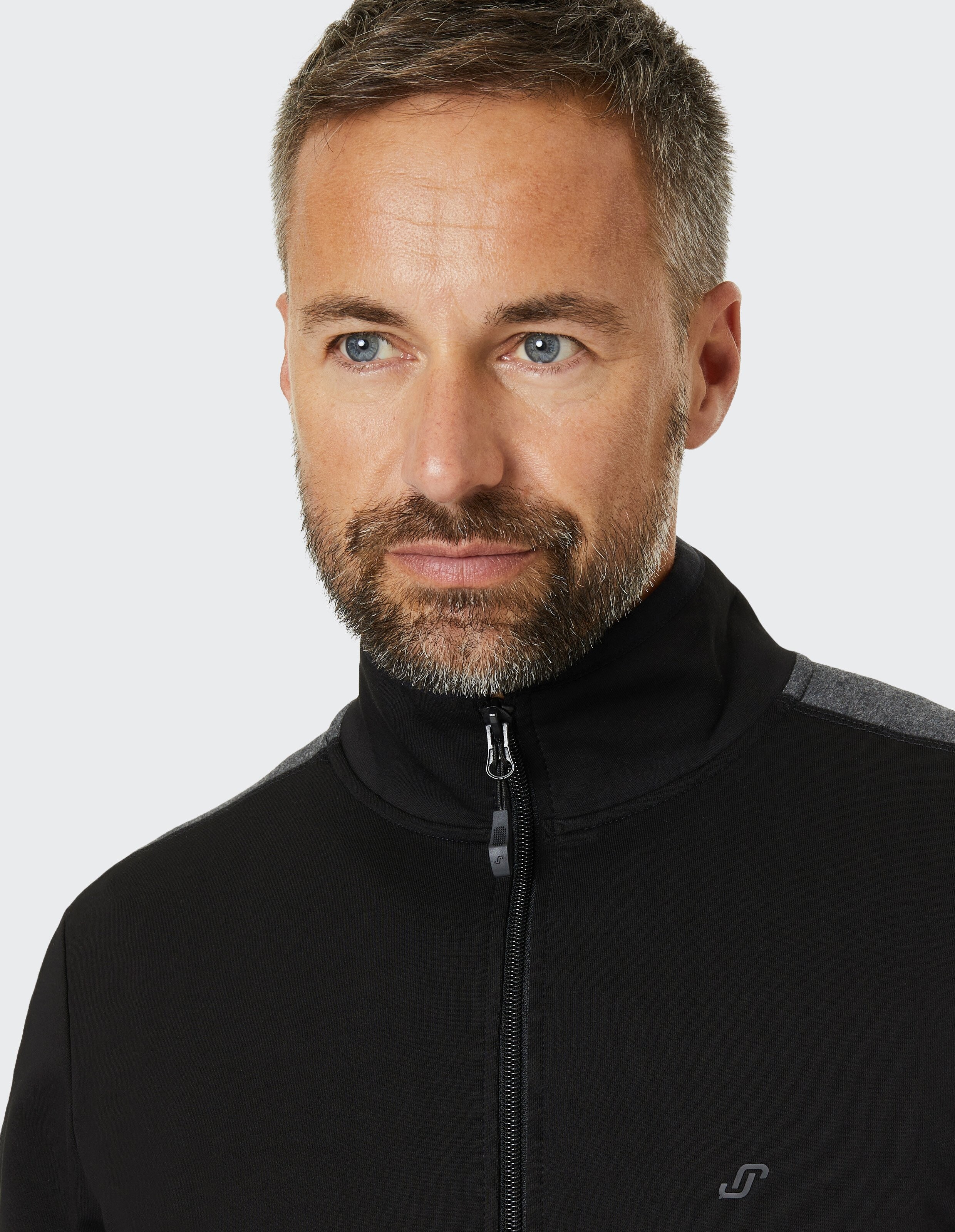Ein Mann mit kurzem Bart und grauem Haar trägt einen schwarzen Rollkragenpullover vor einem neutralen Hintergrund.