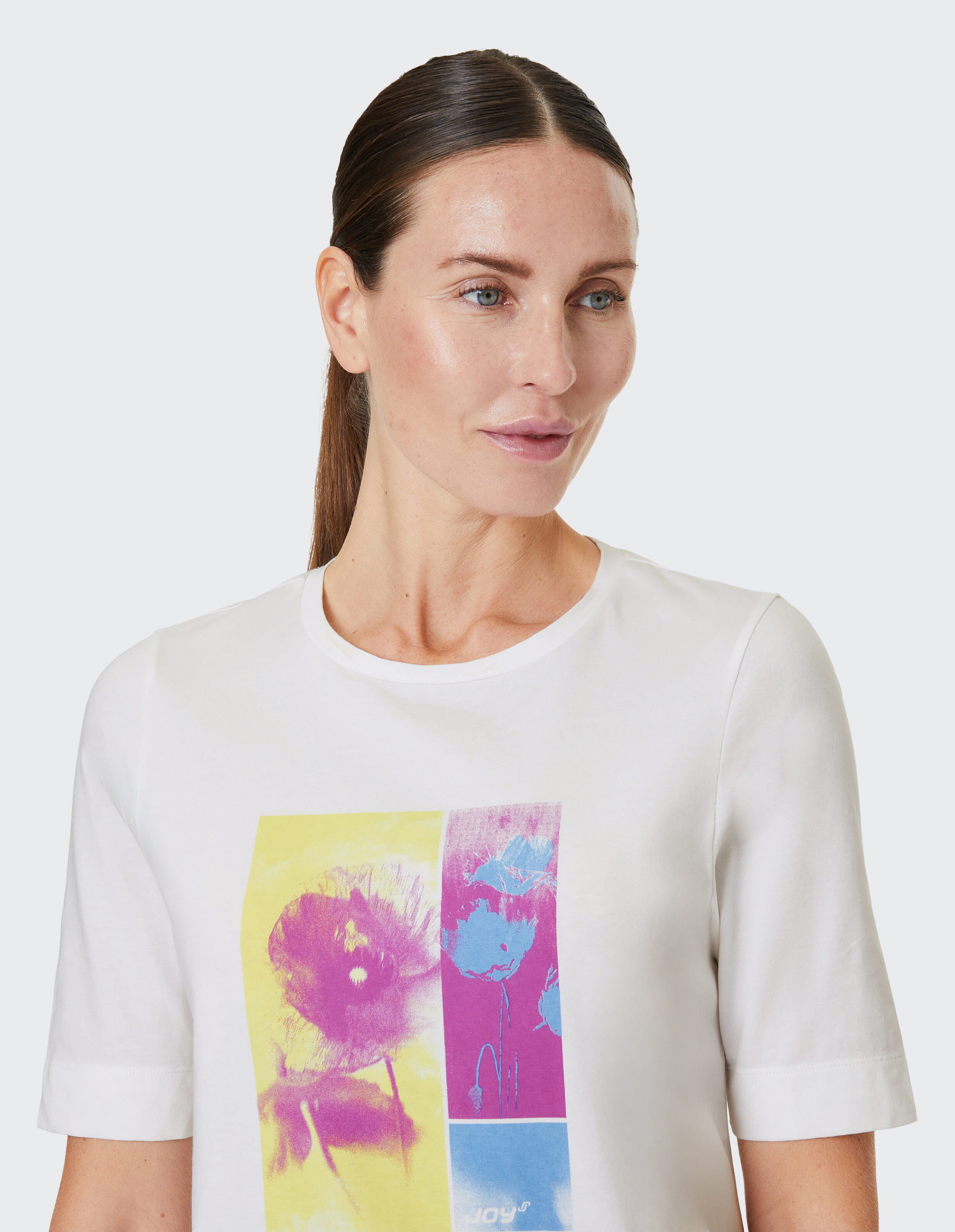 Eine Frau mit zurückgebundenem Haar trägt ein weißes T-Shirt mit buntem Blumenprint in Gelb, Pink und Blau auf der Vorderseite.