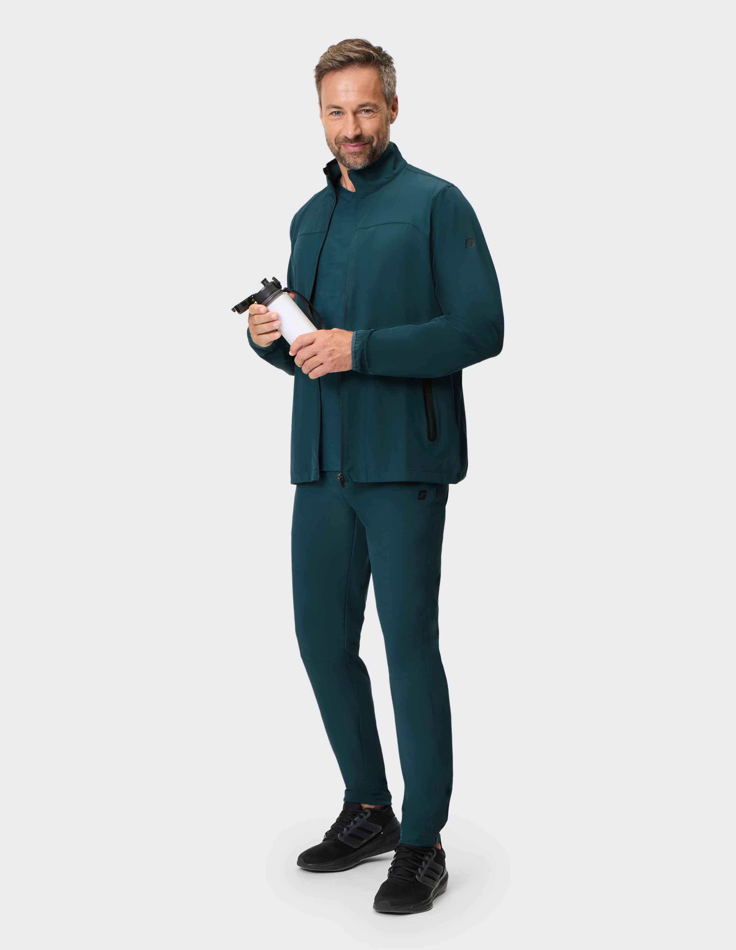 Mann trägt eine dunkelgrüne, lässige Jacke mit Reißverschluss und eine passende Hose und hält eine Wasserflasche in der Hand.