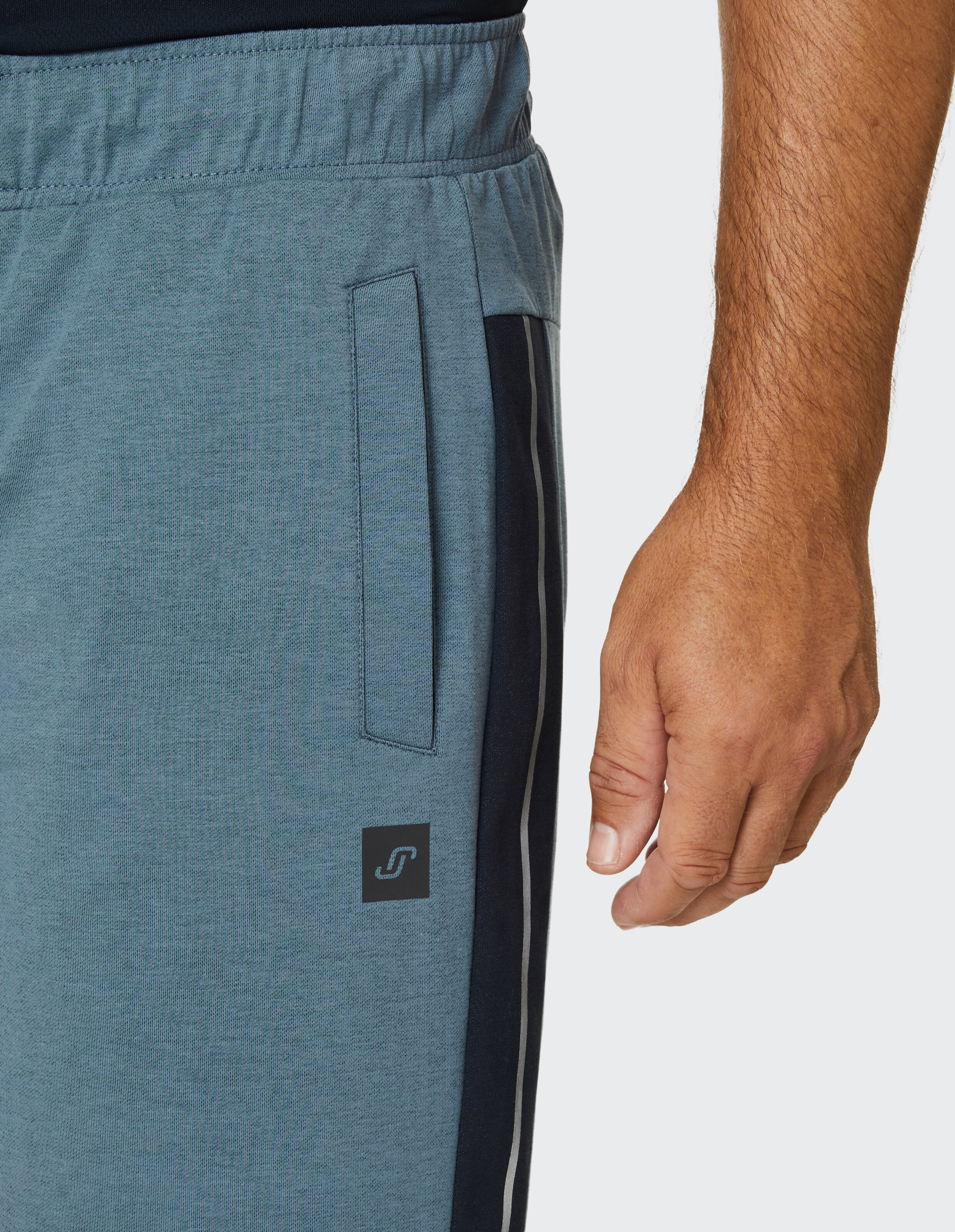 Ein Mann trägt eine blaue Jogginghose mit seitlicher Tasche und Streifen, sein Arm ist entspannt an seiner Seite herunterhängend.