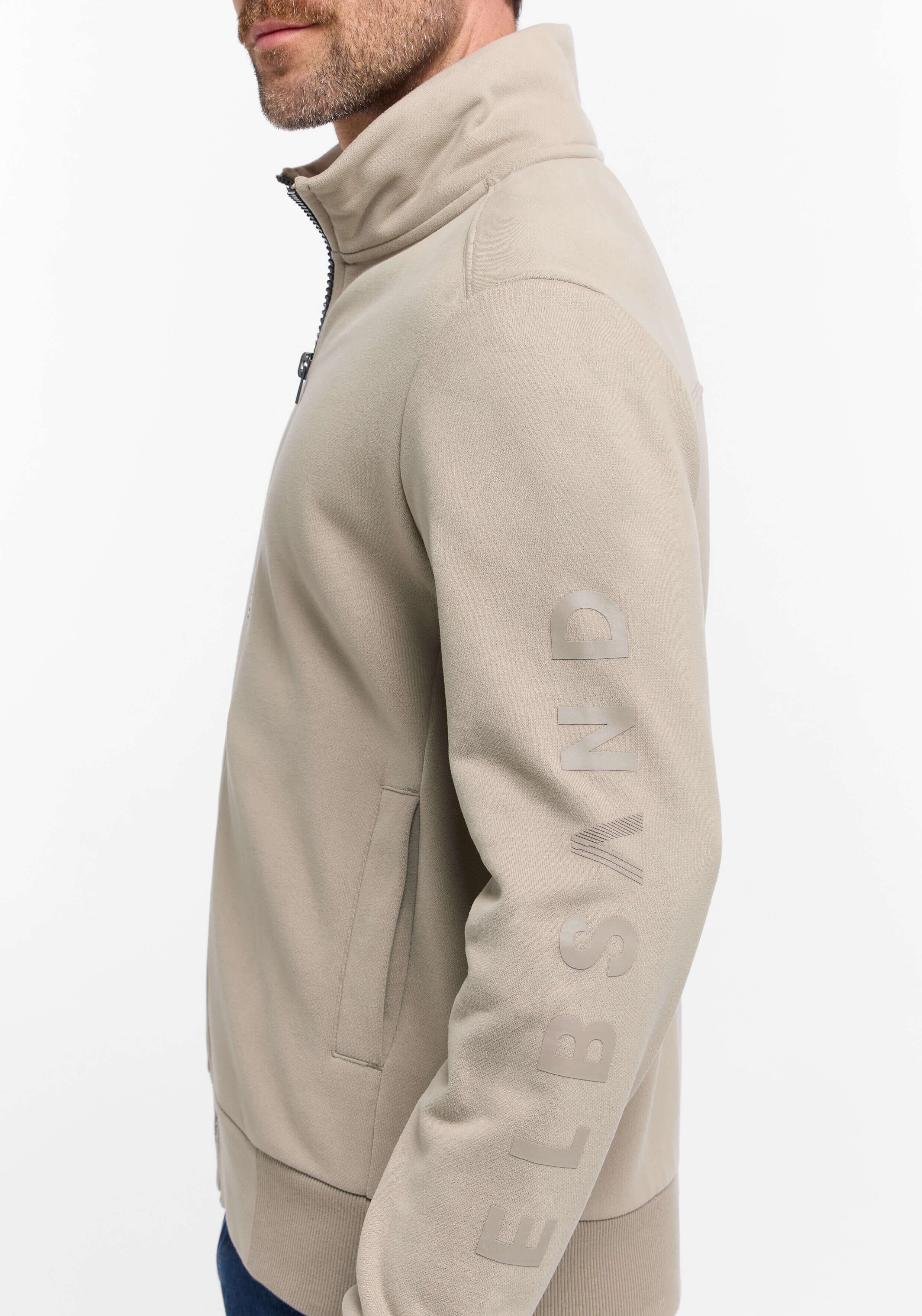 Mann trägt ein beiges ELBSAND-Sweatshirt mit Reißverschluss, hohem Kragen und Logo am Ärmel.