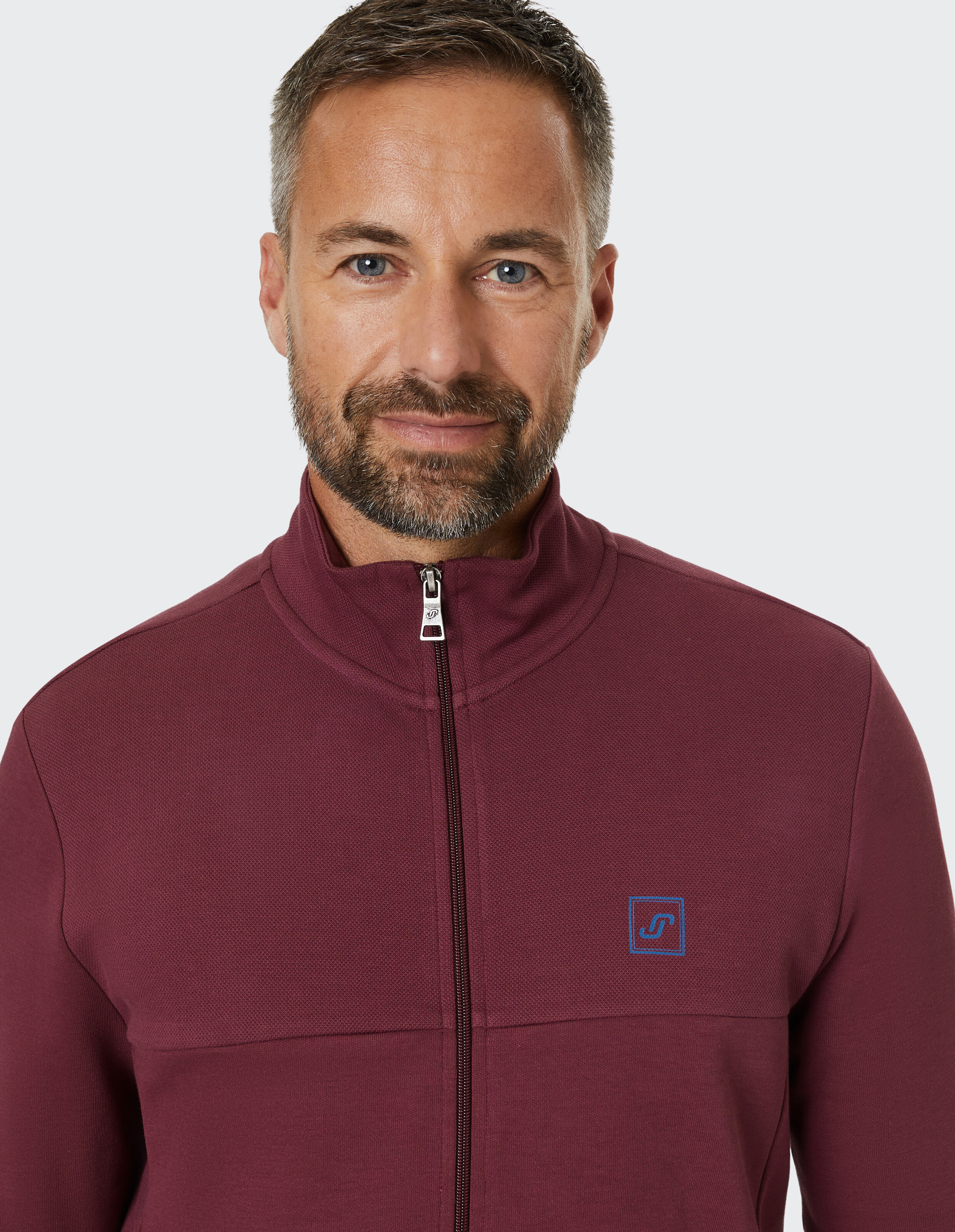 Mann mit braunen Haaren trägt einen dunkelroten Reißverschluss-Pullover mit kleinem blauen Logo auf hellem Hintergrund.