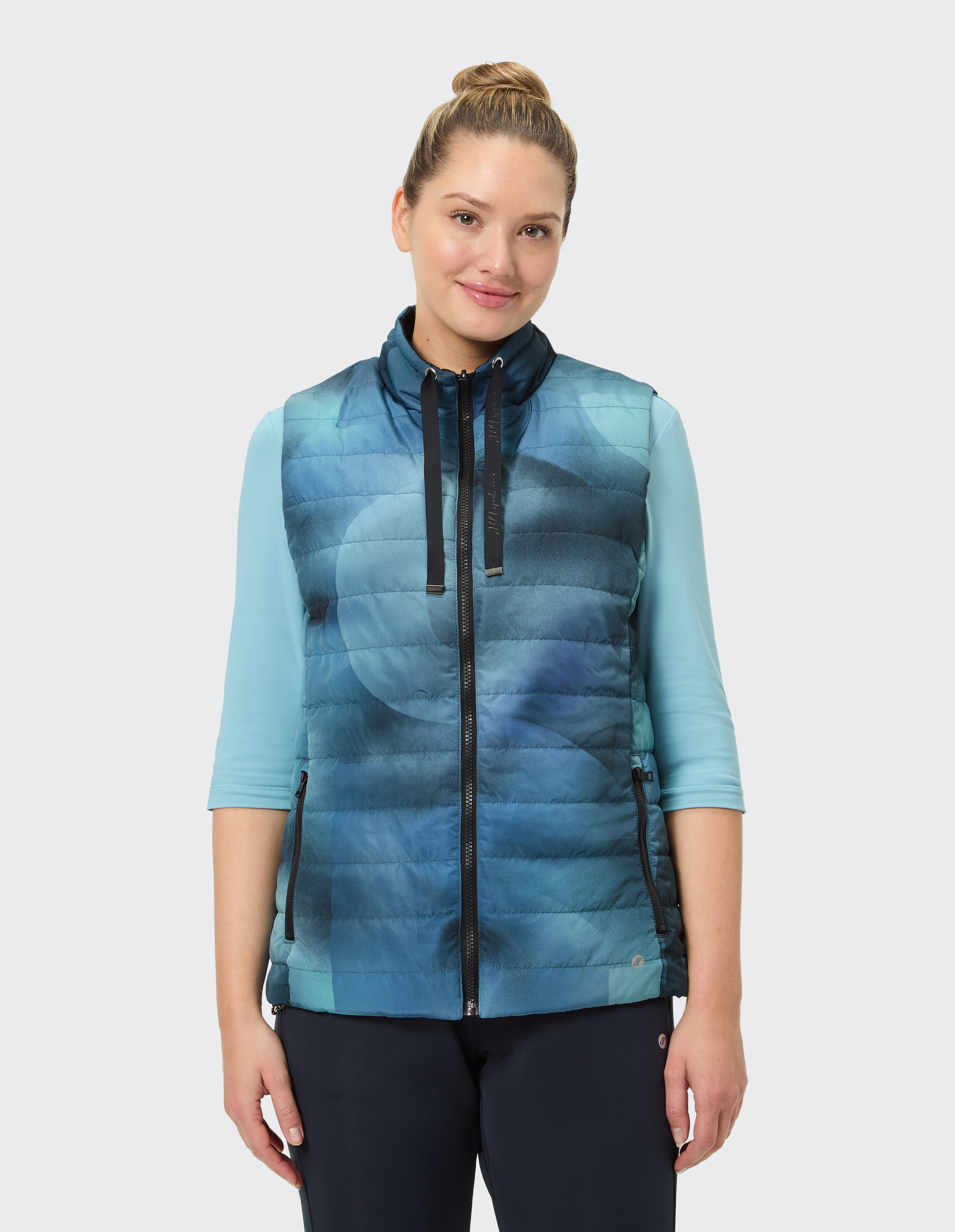 Eine Frau trägt eine blau gemusterte Steppweste über einem hellblauen Shirt mit Dreiviertelärmeln, vor weißem Hintergrund.