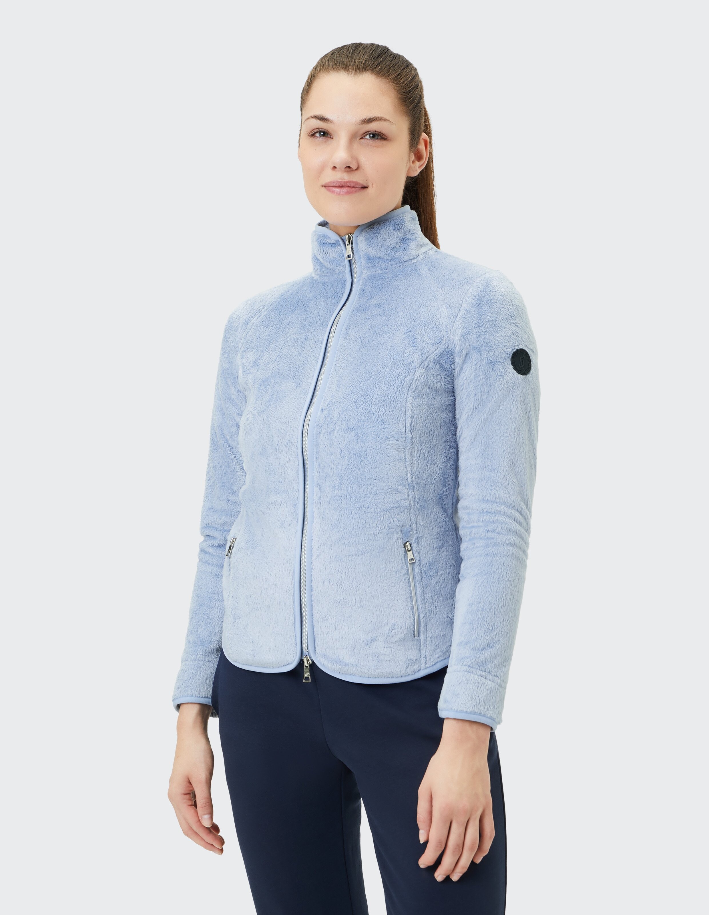 Frau trägt eine flauschige, hellblaue Jacke mit Reißverschluss und hohem Kragen, kombiniert mit einer dunkelblauen Hose.