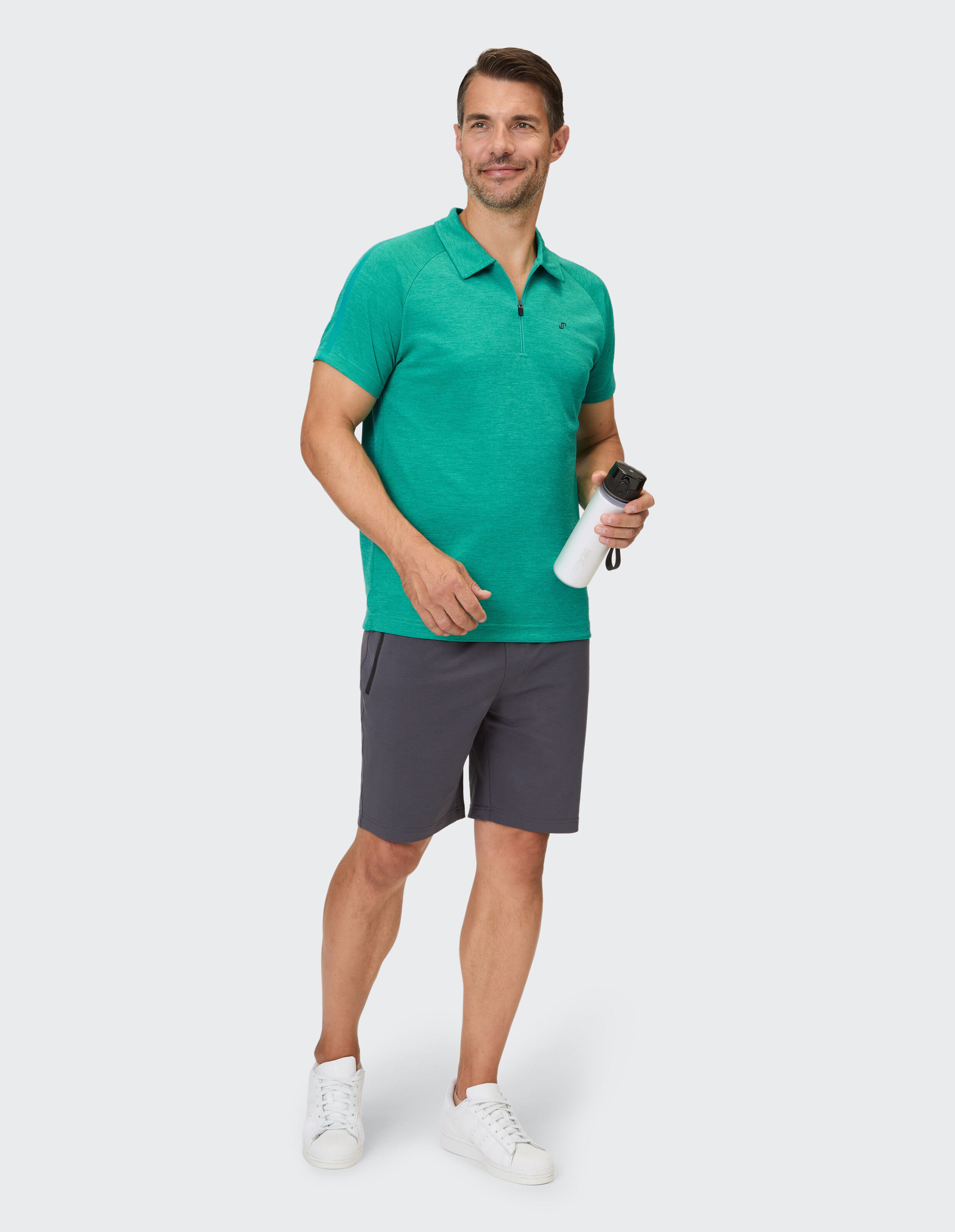 Ein Mann trägt ein kurzärmeliges, smaragdgrünes Poloshirt mit halblangem Reißverschluss und dunkelgraue Shorts, dazu weiße Sneaker.