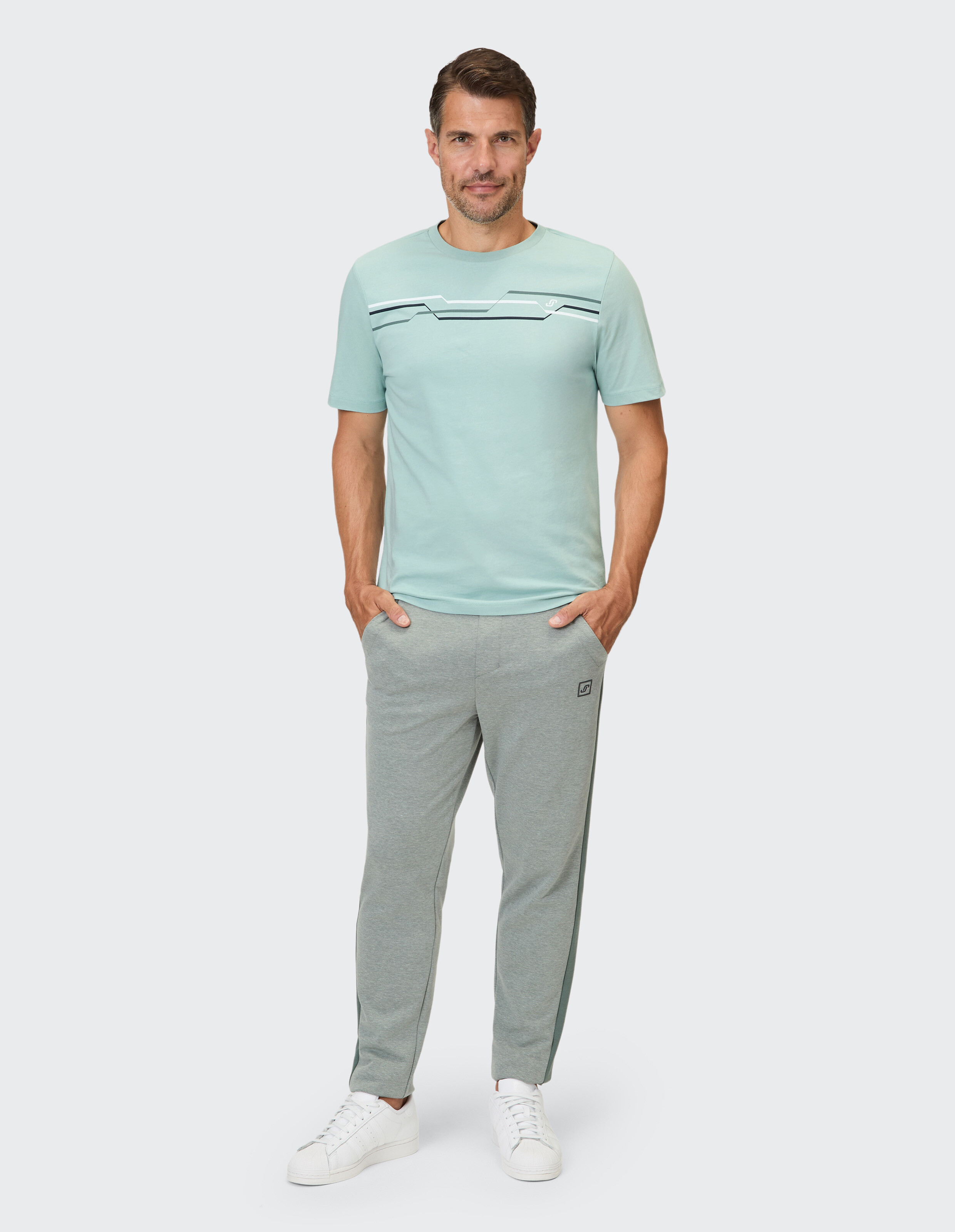 Ein Mann trägt ein mintgrünes T-Shirt mit horizontalen Streifen und eine graue Jogginghose mit Seitenstreifen.