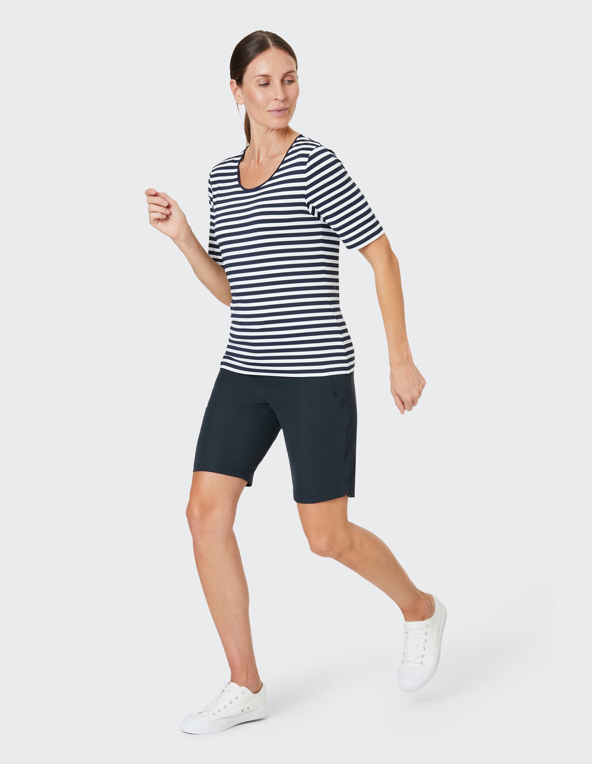 Frau in gestreiftem T-Shirt und dunklen Shorts geht sportlich, trägt weiße Turnschuhe vor schlichtem Hintergrund.