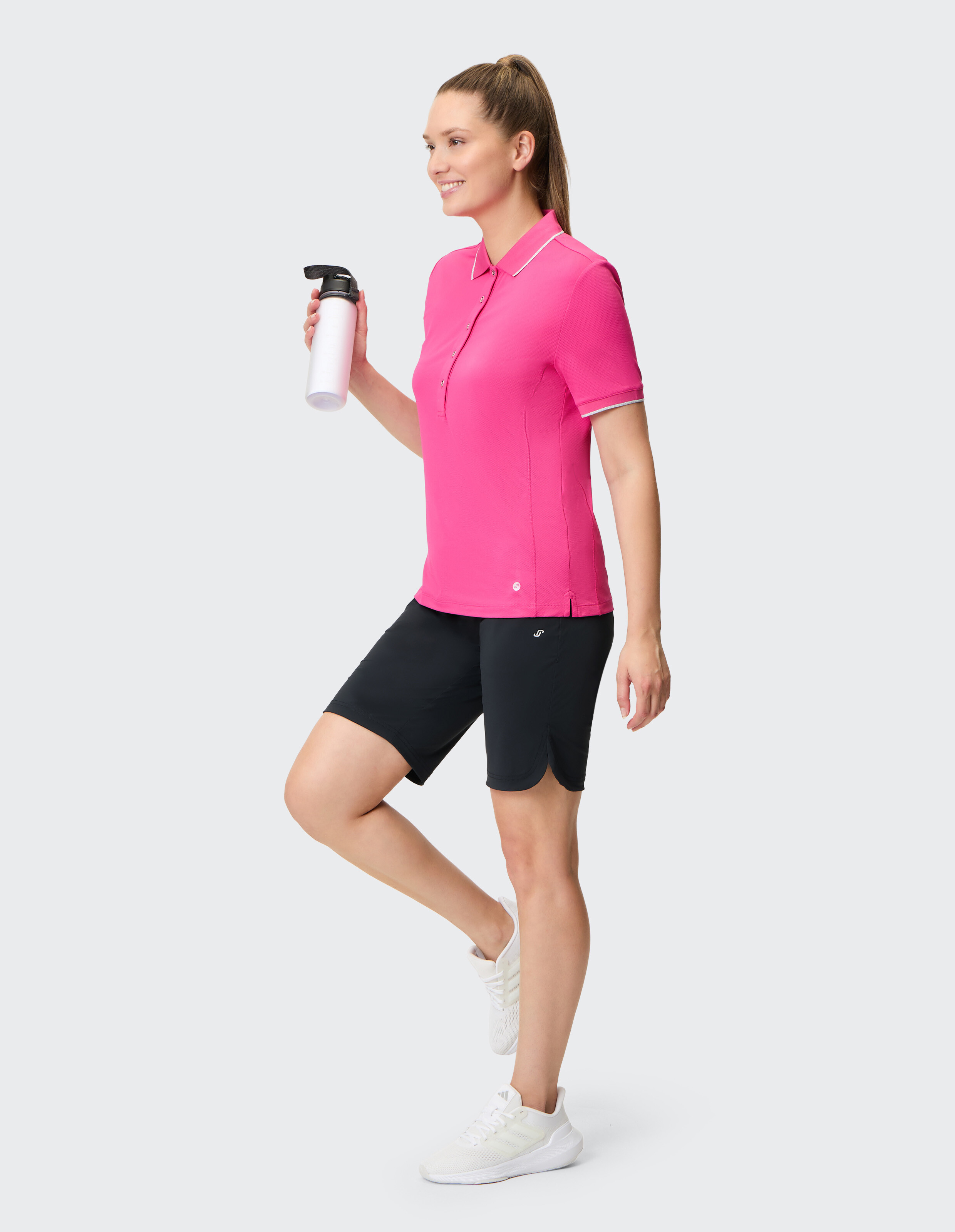 Frau in einem rosa Poloshirt und schwarzen Shorts mit einer Wasserflasche für lässige Aktivkleidung.