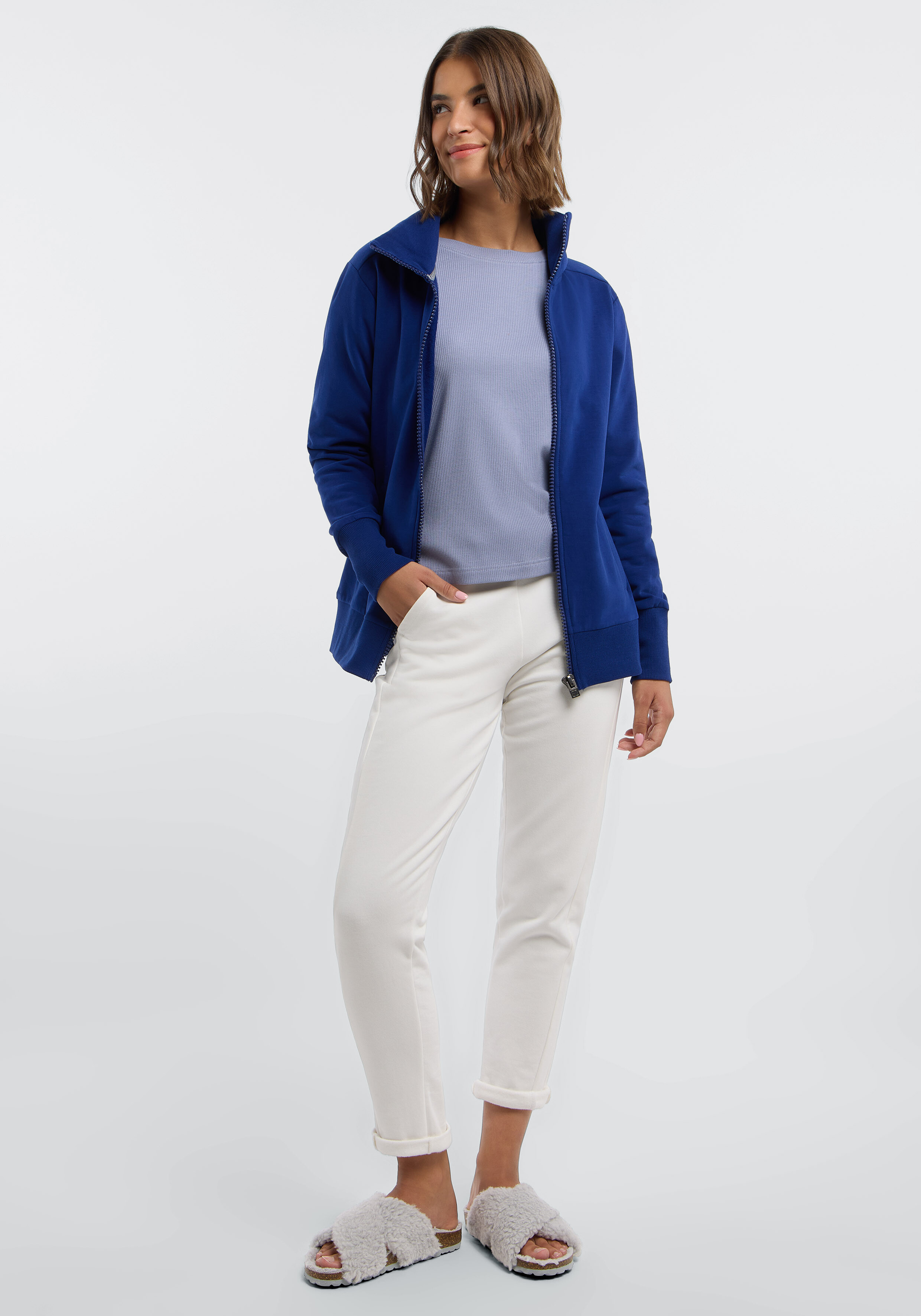 Frau trägt eine blaue Jacke mit Reißverschluss, ein helles Oberteil und eine weiße Freizeithose.