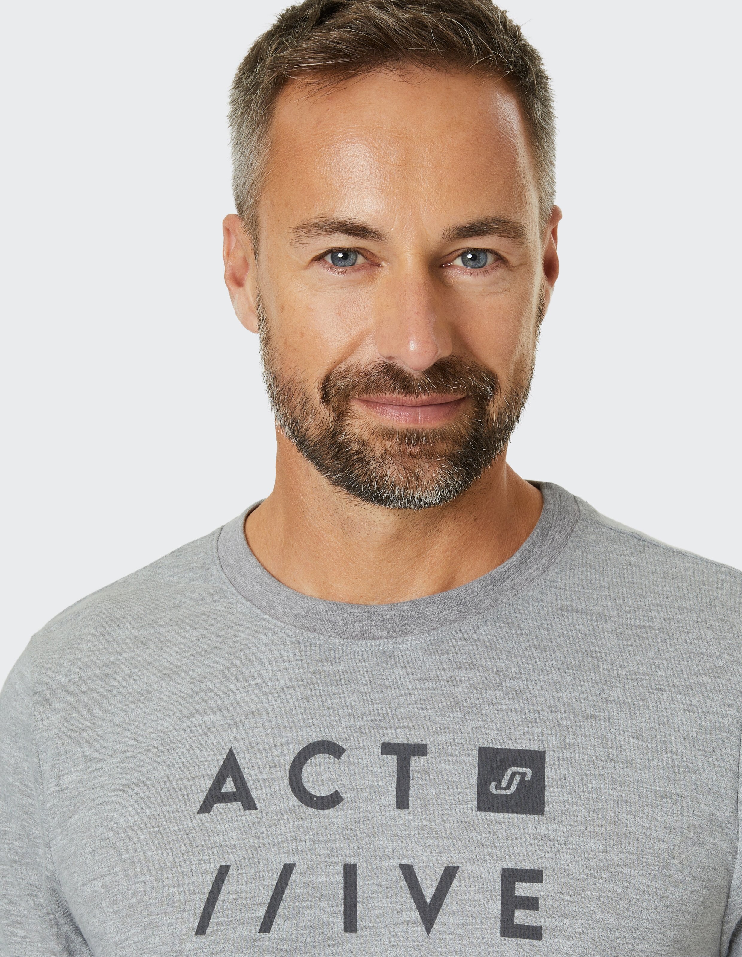 Ein Mann mit grauem Bart und kurzen Haaren trägt ein graues Shirt mit der Aufschrift "ACT /// IVE", lächelnd vor neutralem Hintergrund.