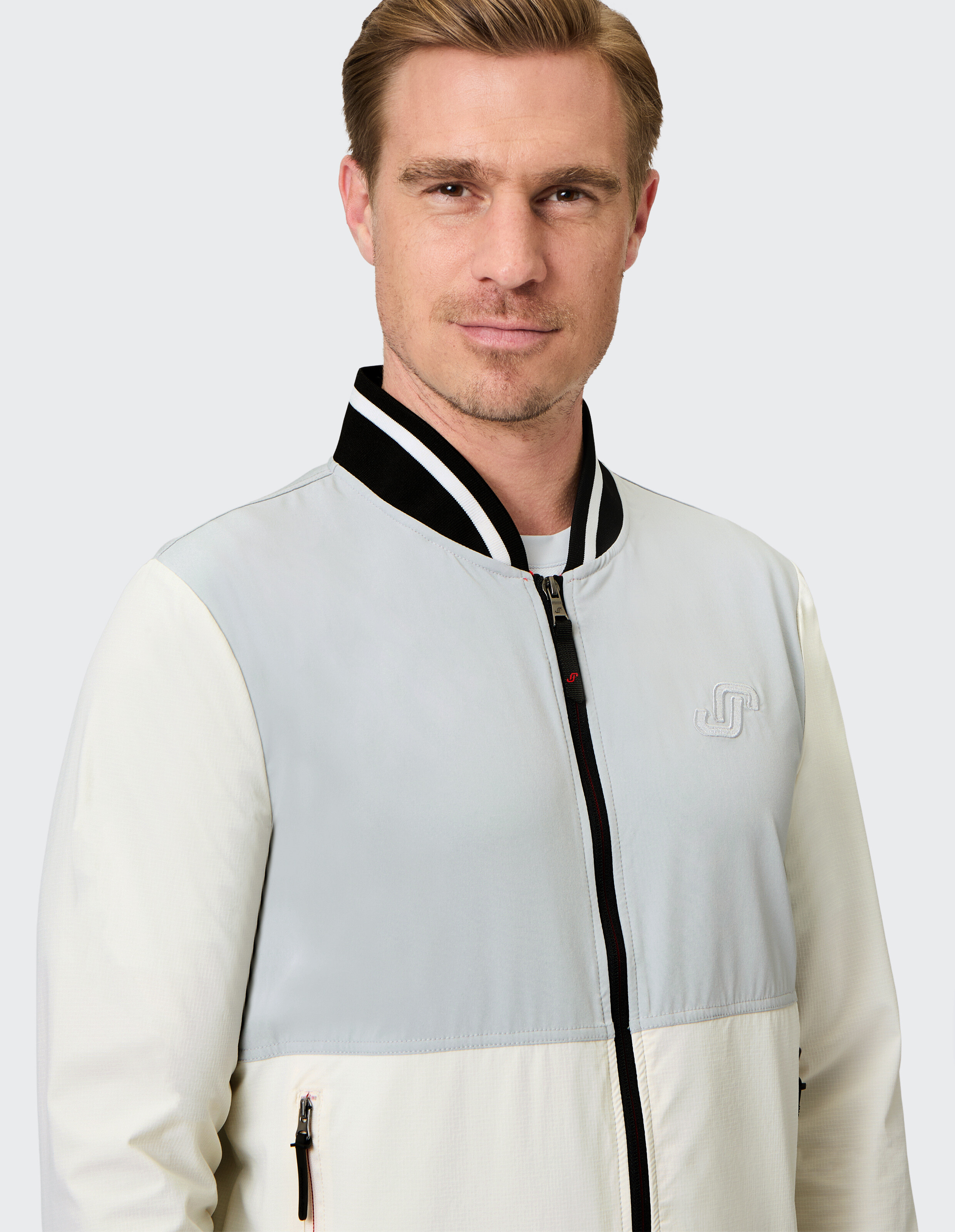 Ein Mann trägt eine Jacke mit hellem Oberteil, dunklem Kragen und Reißverschluss; er schaut direkt in die Kamera.