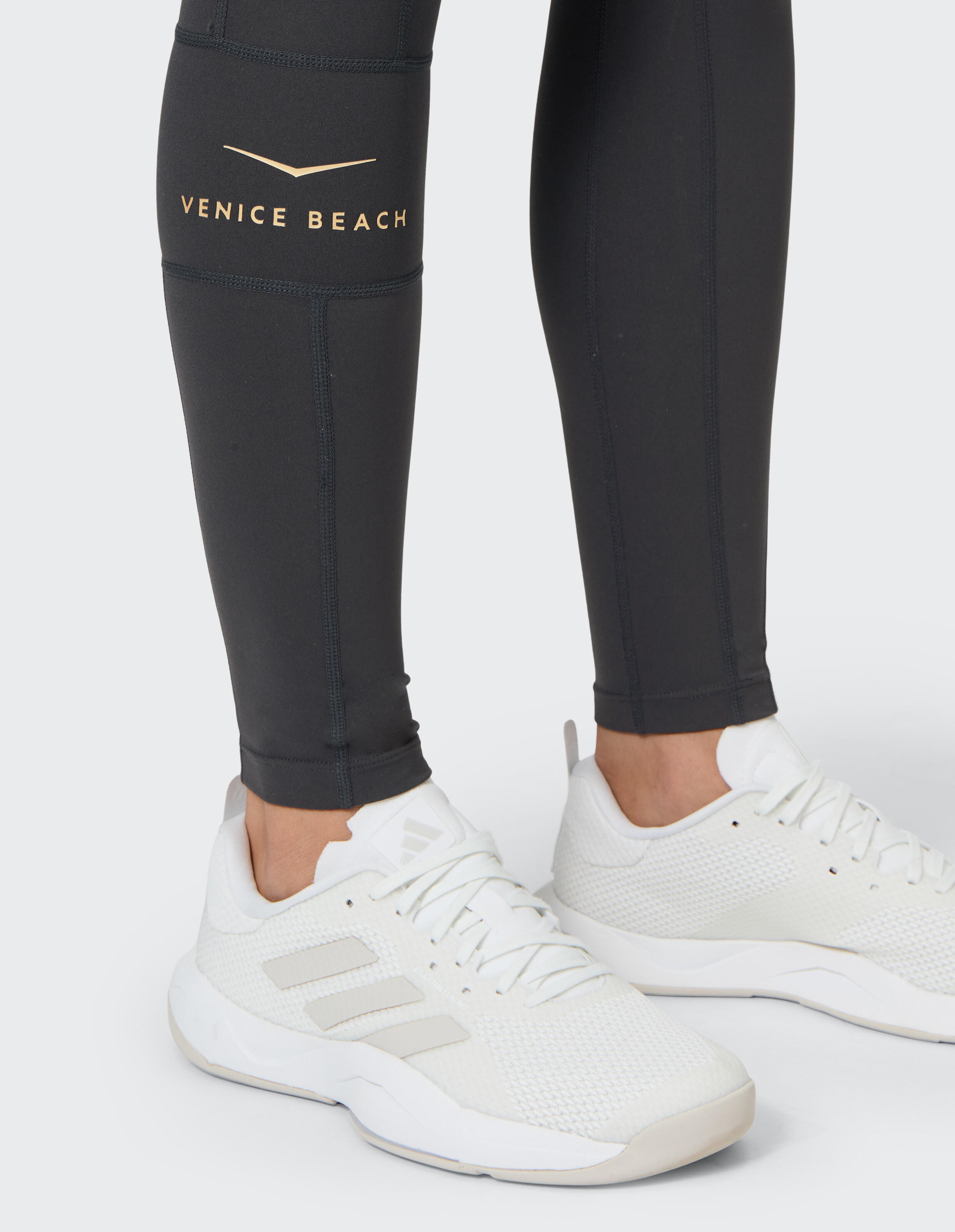 Nahaufnahme einer schwarzen Leggings von Venice Beach mit goldenem Logo und weißen Sportschuhen.