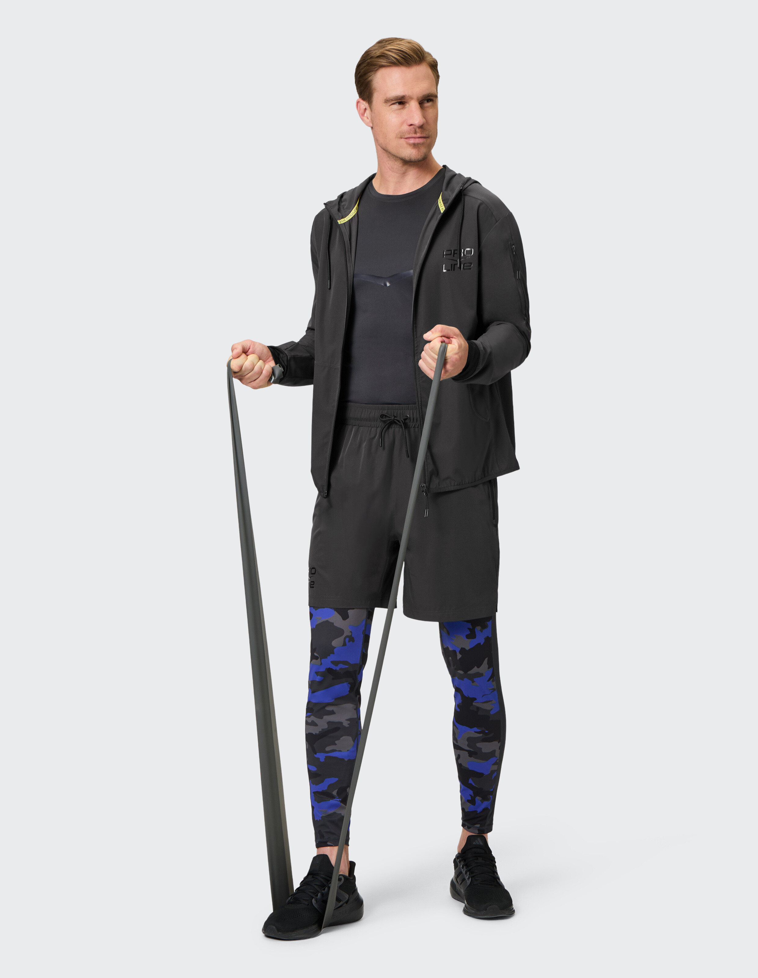 Mann in lässiger Sportkleidung mit schwarzer Jacke, blauen Camo-Leggings und Widerstandsbändern.