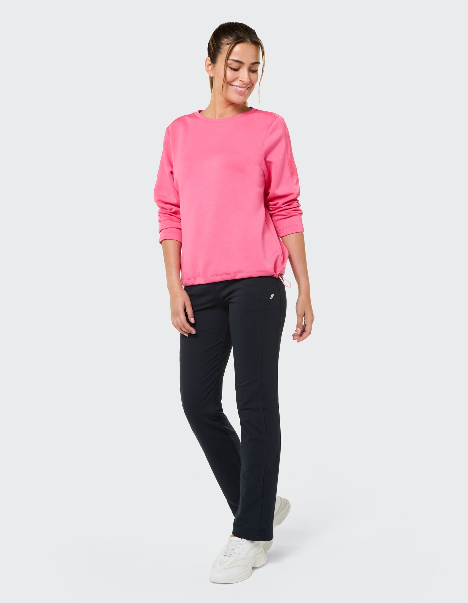 Frau in rosa Sweatshirt und schwarzer Hose, lächelnd in Freizeitkleidung.
