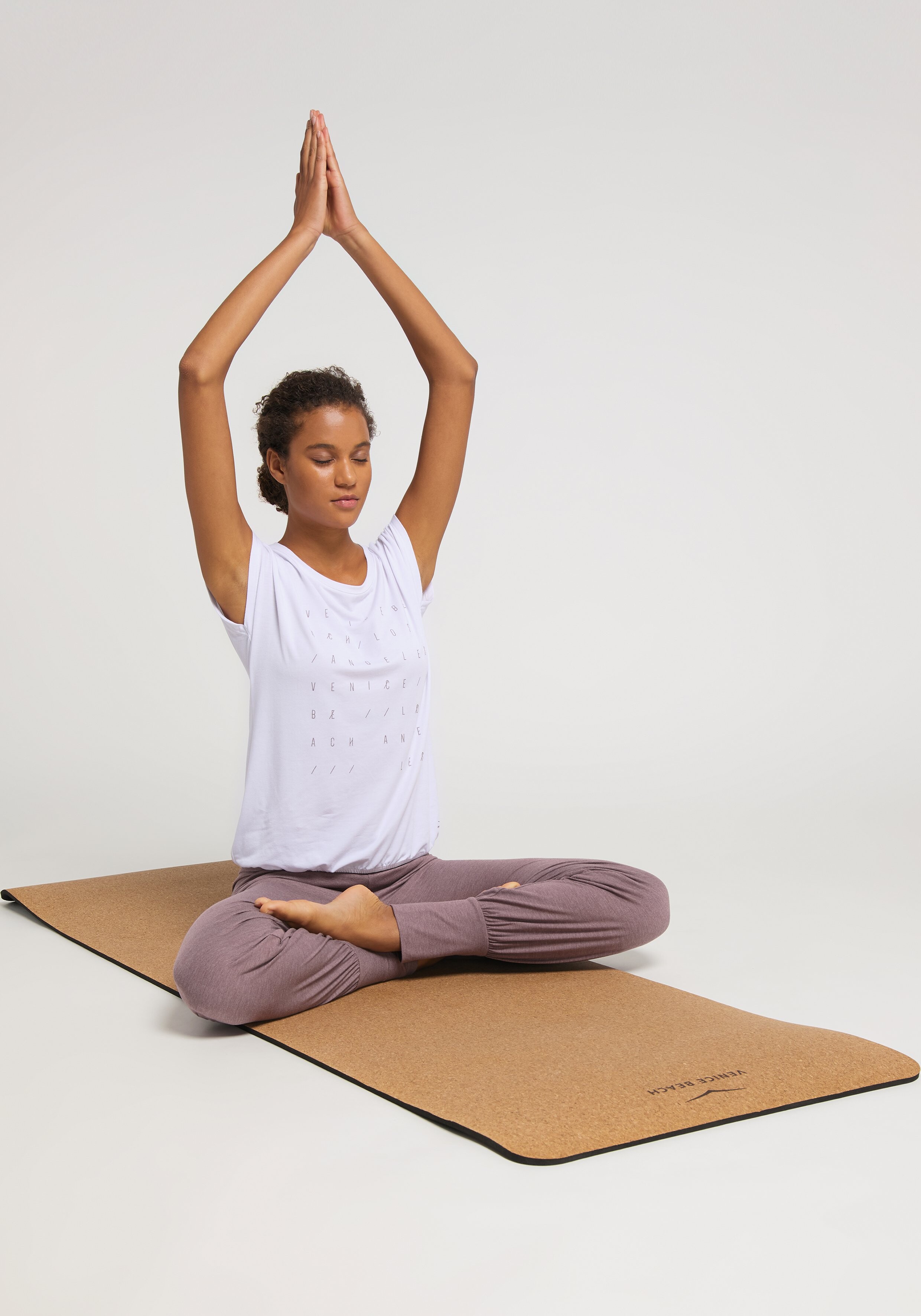 Frau in einem weißen Top und malvenfarbenen Jogginghosen macht Yoga auf einer Korkmatte.
