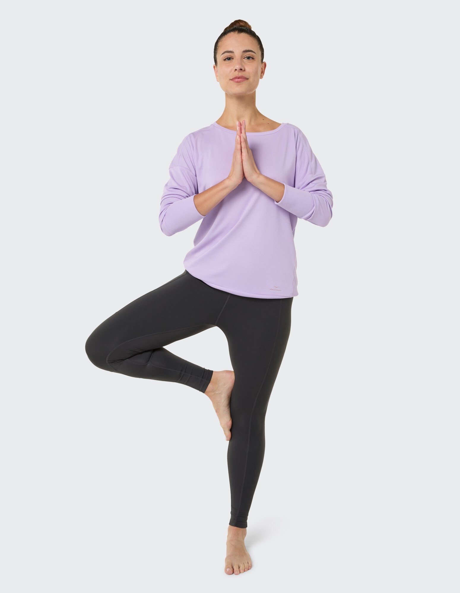 Frau in einem lavendelfarbenen Langarmshirt und schwarzen Leggings praktiziert Yoga.