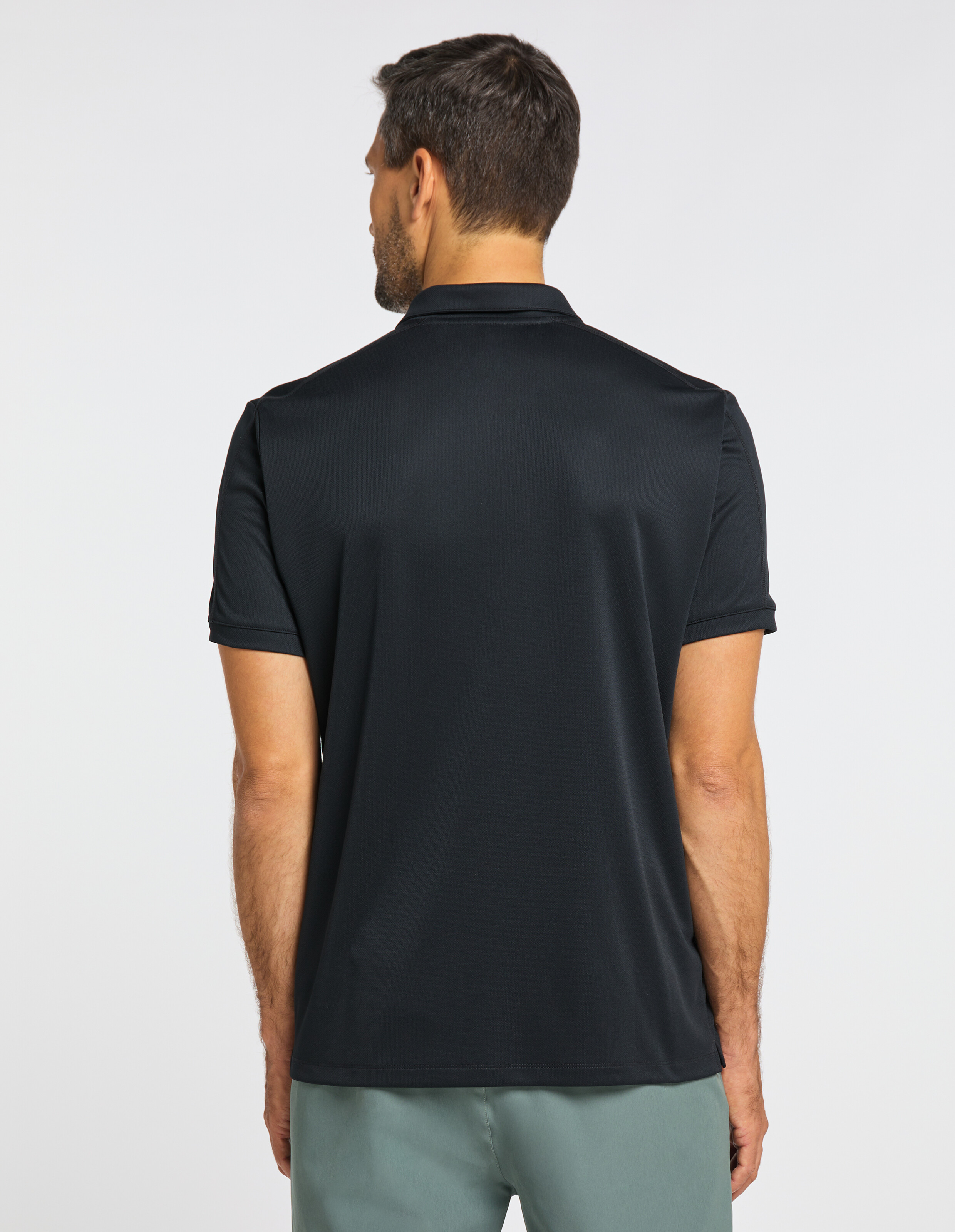 Mann mit dunklem, kurzärmligem Polohemd und grauer Hose steht von hinten sichtbar vor weißem Hintergrund.