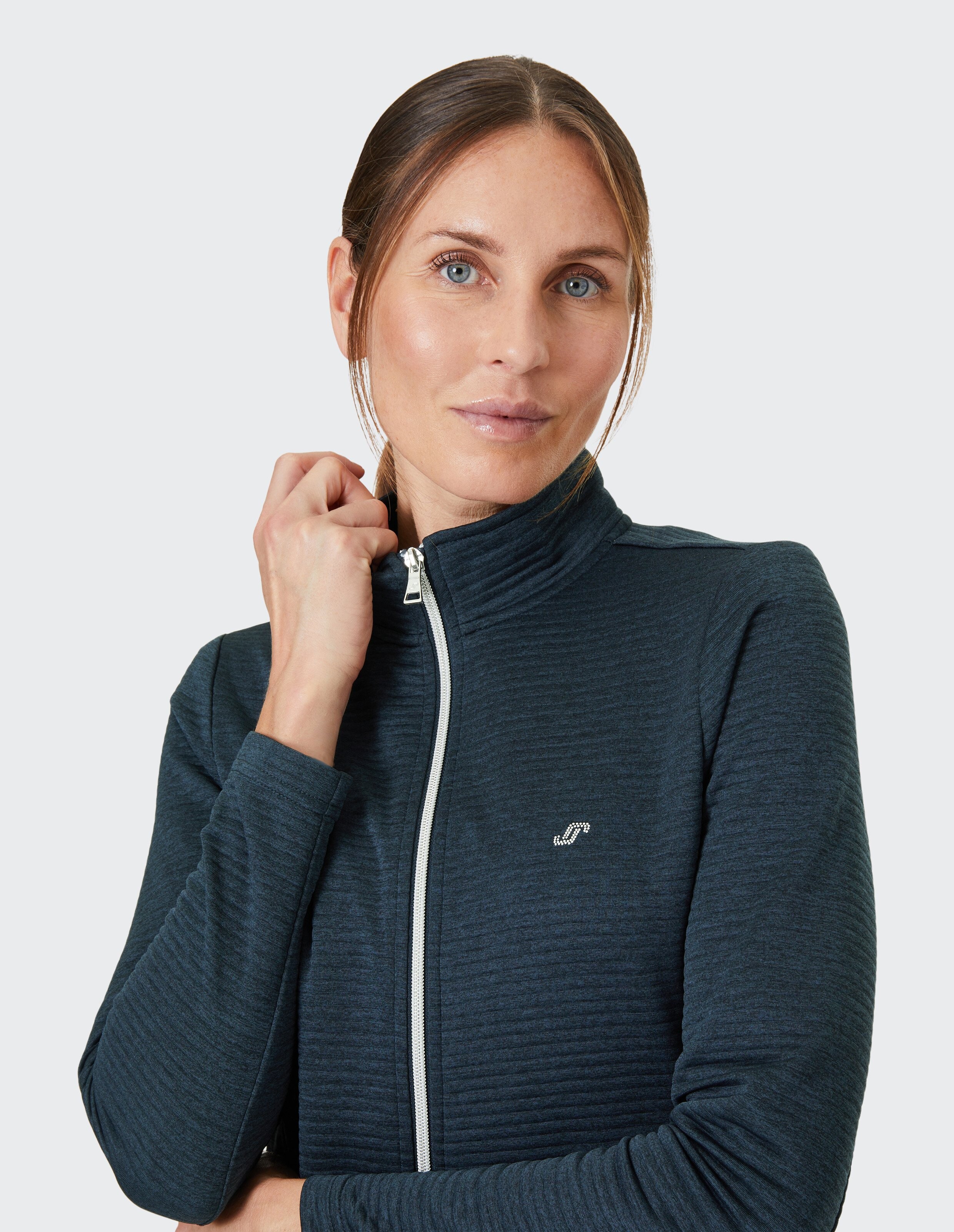 Frau mit hellem Teint und blauen Augen in einer dunkelblauen, strukturierten Jacke mit Reißverschluss vor grauem Hintergrund.
