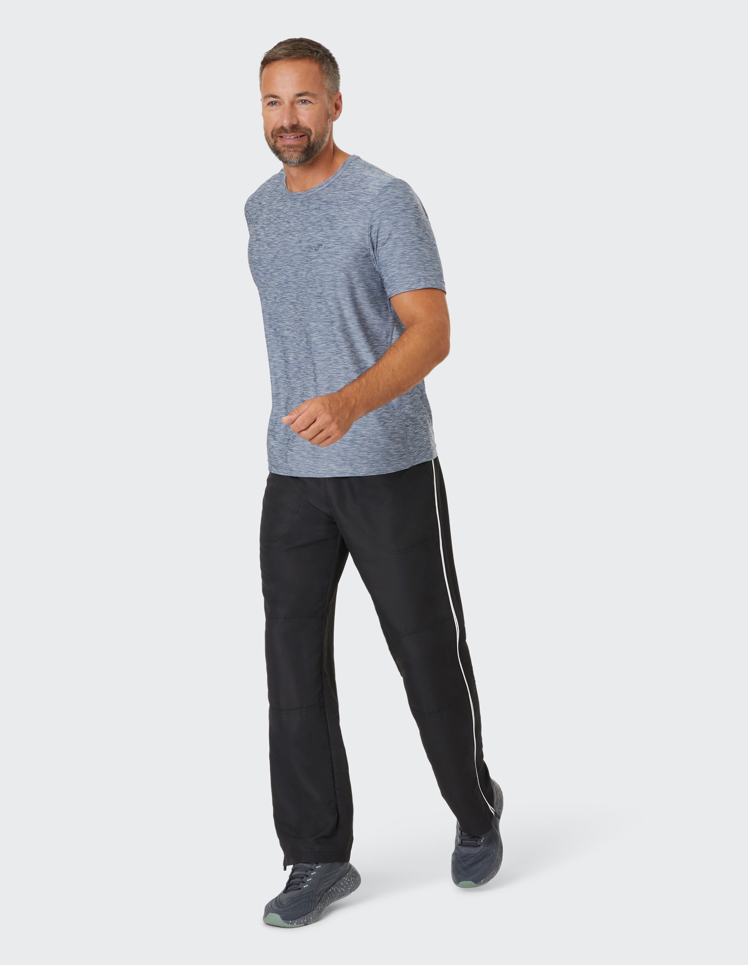 Mann mit blauem T-Shirt und schwarzer Hose mit Streifen läuft, lächelt und schaut zur Seite; hellgrauer Hintergrund.
