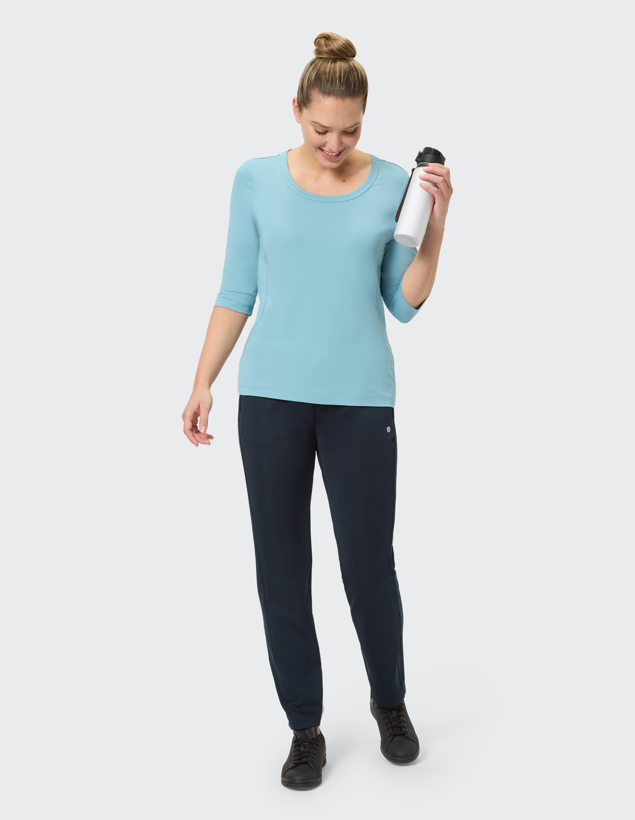Frau in hellblauem Oberteil und dunkelblauer Hose, mit einer Wasserflasche in der Hand, lässiger Stil.