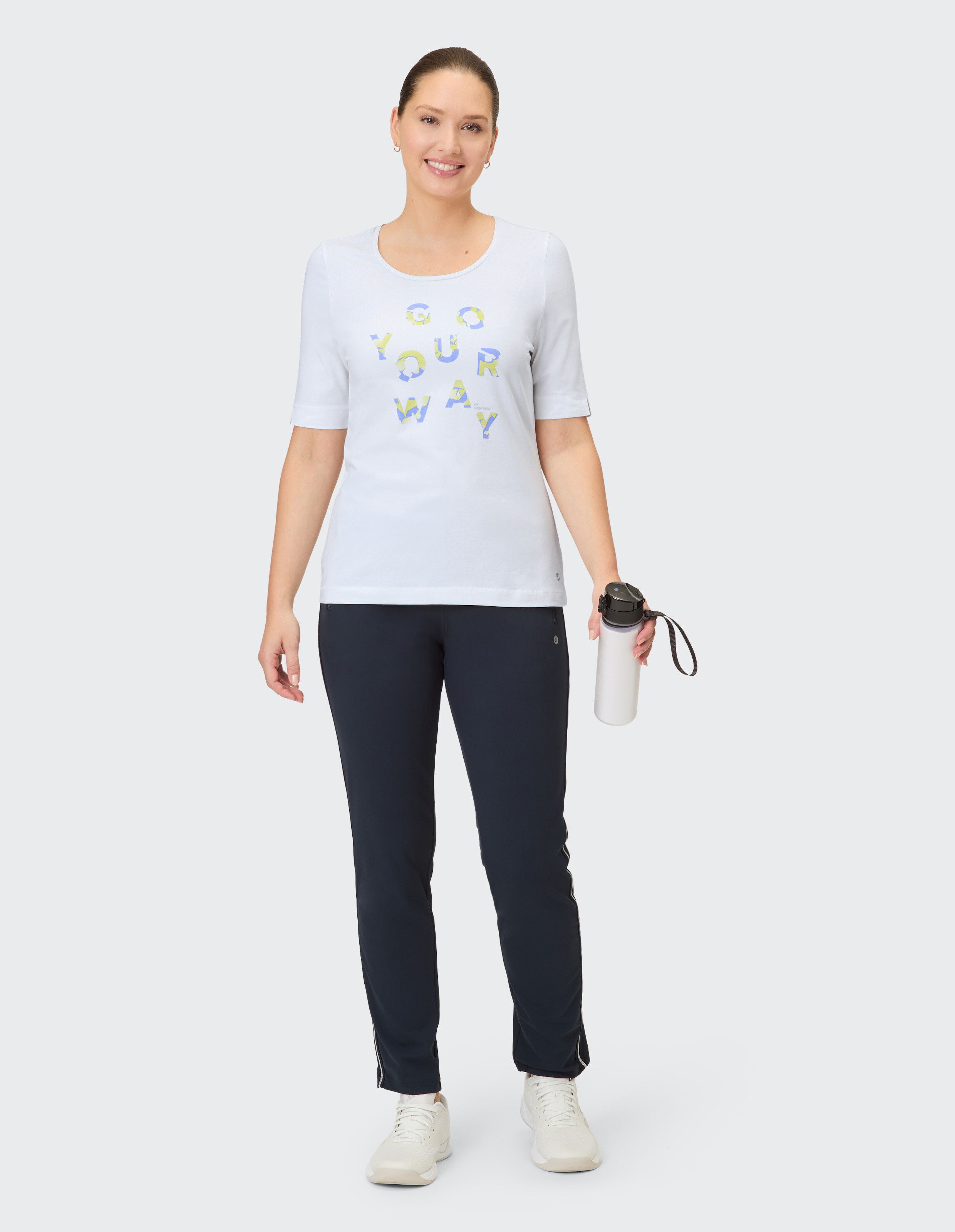 Eine lächelnde Frau in einem weißen Shirt mit blauen und gelben Buchstaben und einer dunklen Hose mit weißem Besatz hält eine weiße Wasserflasche.
