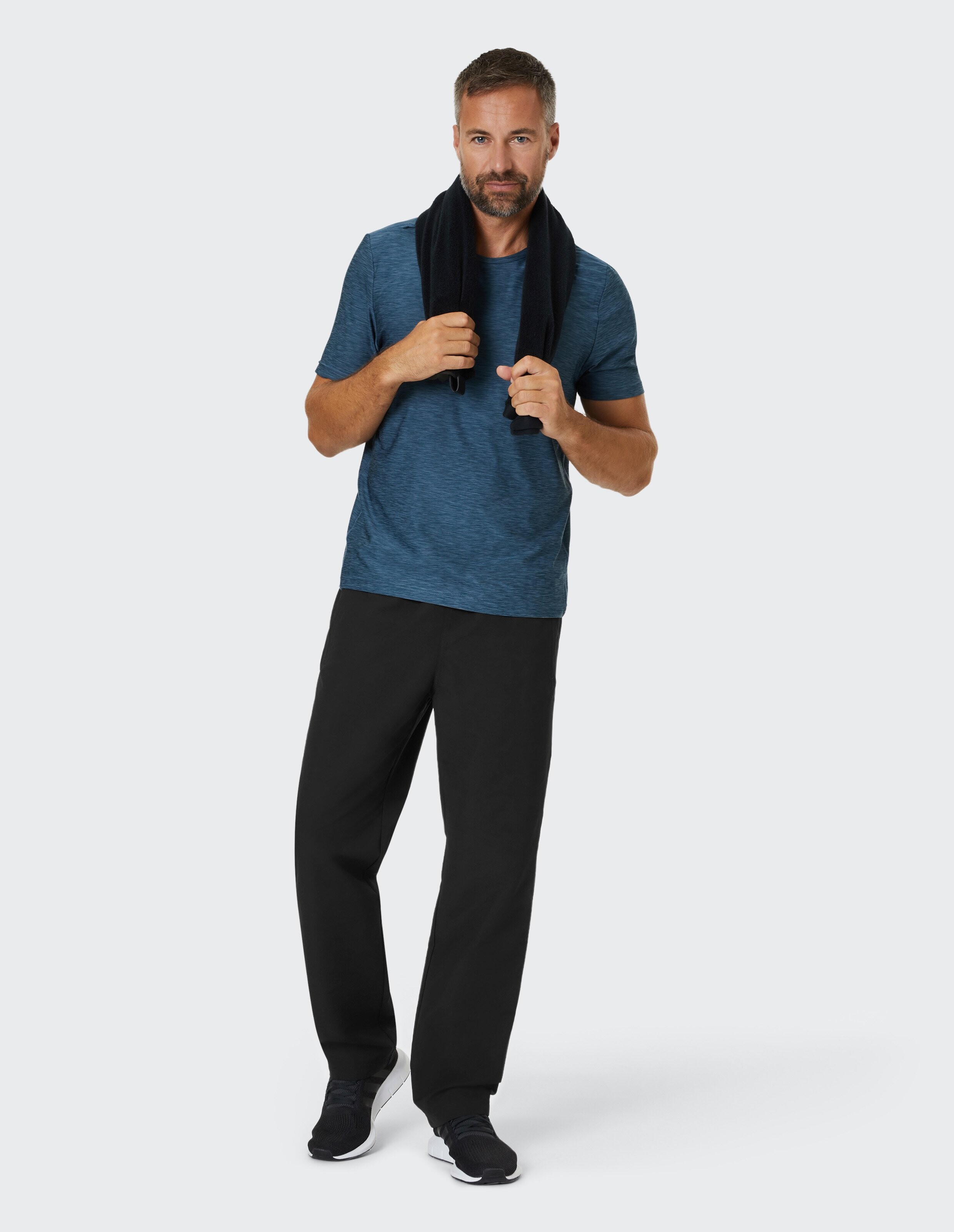 Ein Mann in blauem T-Shirt und schwarzen Hosen hält ein Handtuch um den Nacken, steht lässig und blickt freundlich in die Kamera.