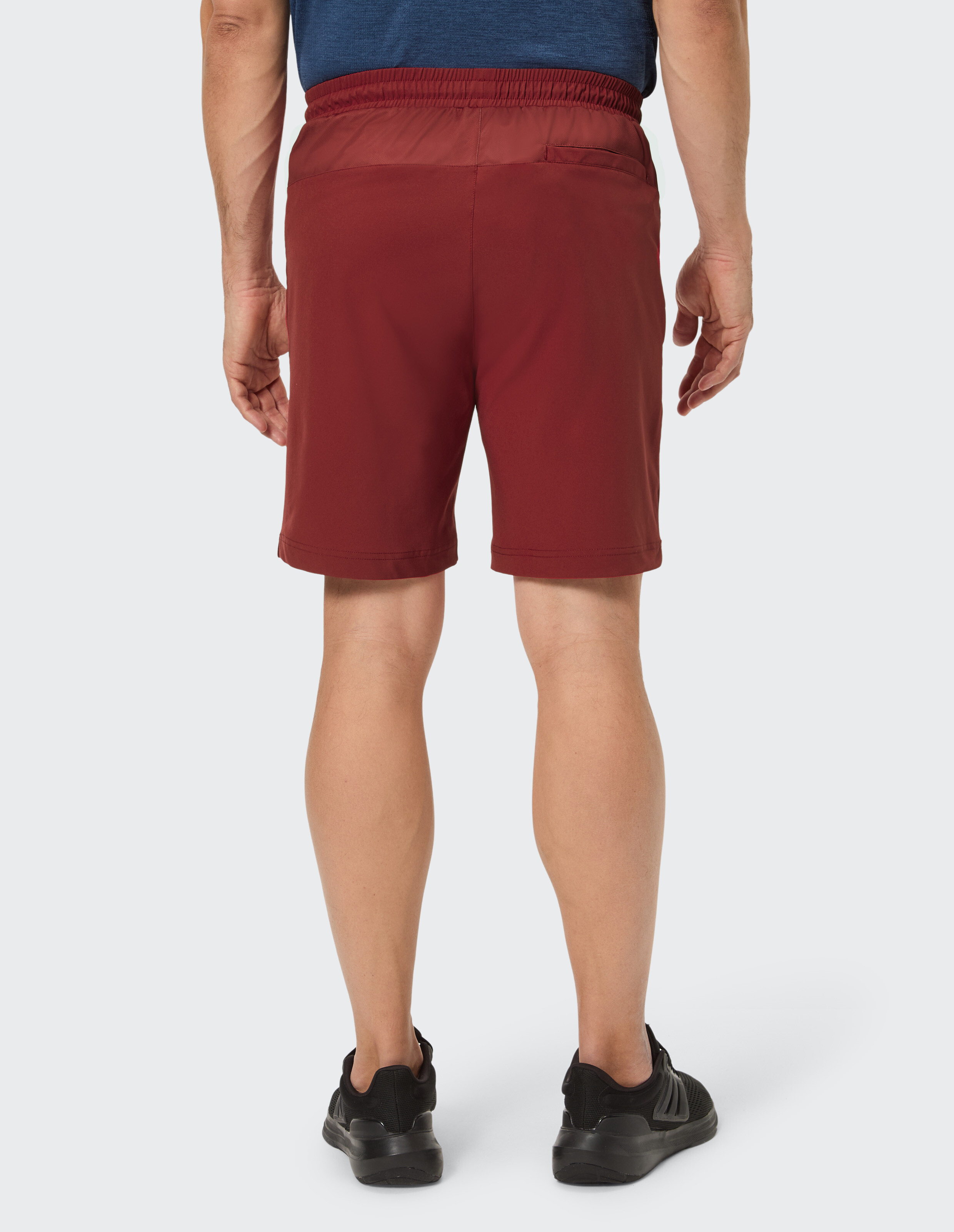 Mann in rotem Shorts und blauem Shirt von hinten, trägt schwarze Schuhe, steht auf hellem Hintergrund, rechte Hand neben der Hüfte.