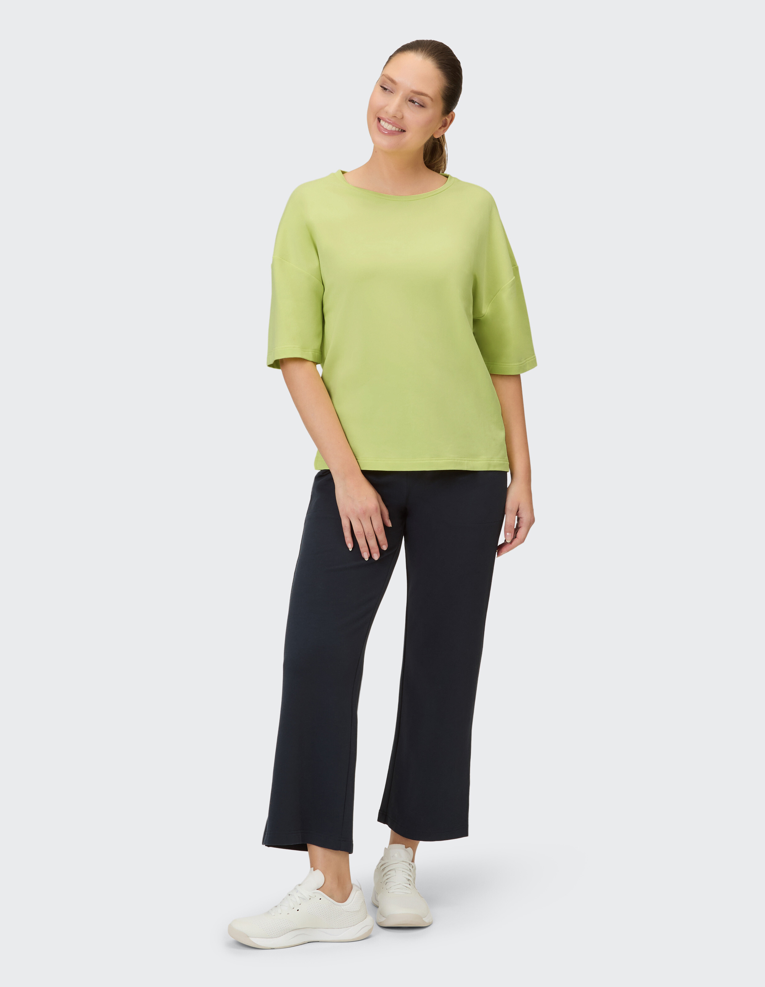 Eine lächelnde Frau trägt ein hellgrünes Kurzarm-Oberteil und eine weite schwarze Hose.