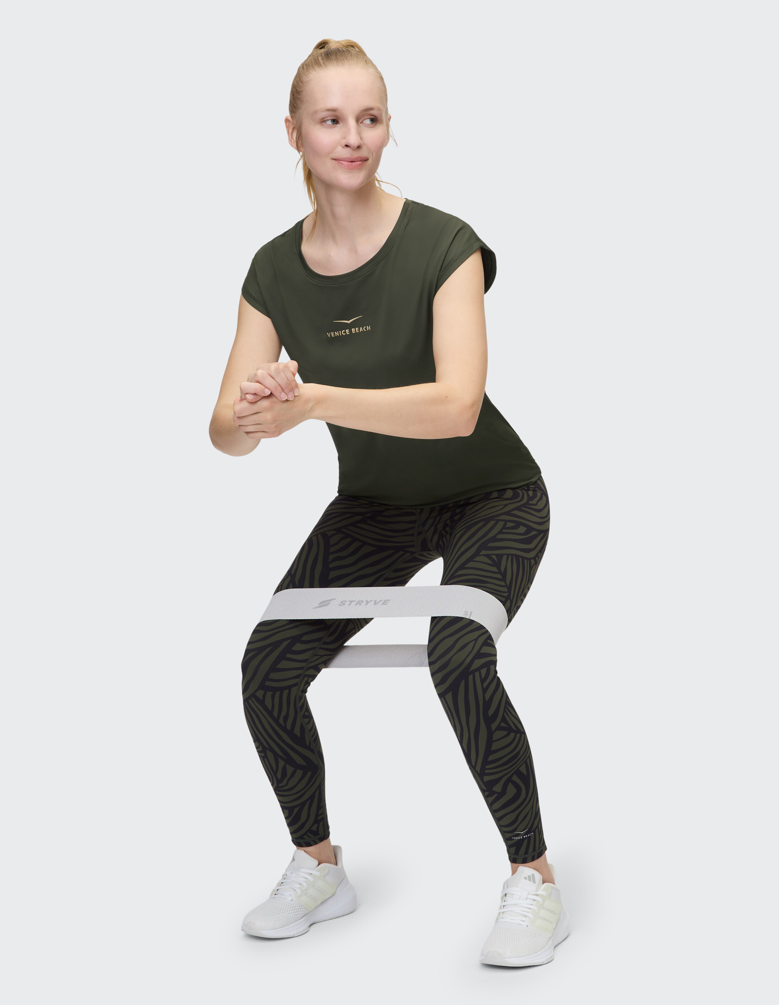 Frau in olivgrünem T-Shirt und gemusterten Leggings macht Kniebeugen mit einem Widerstandsband.