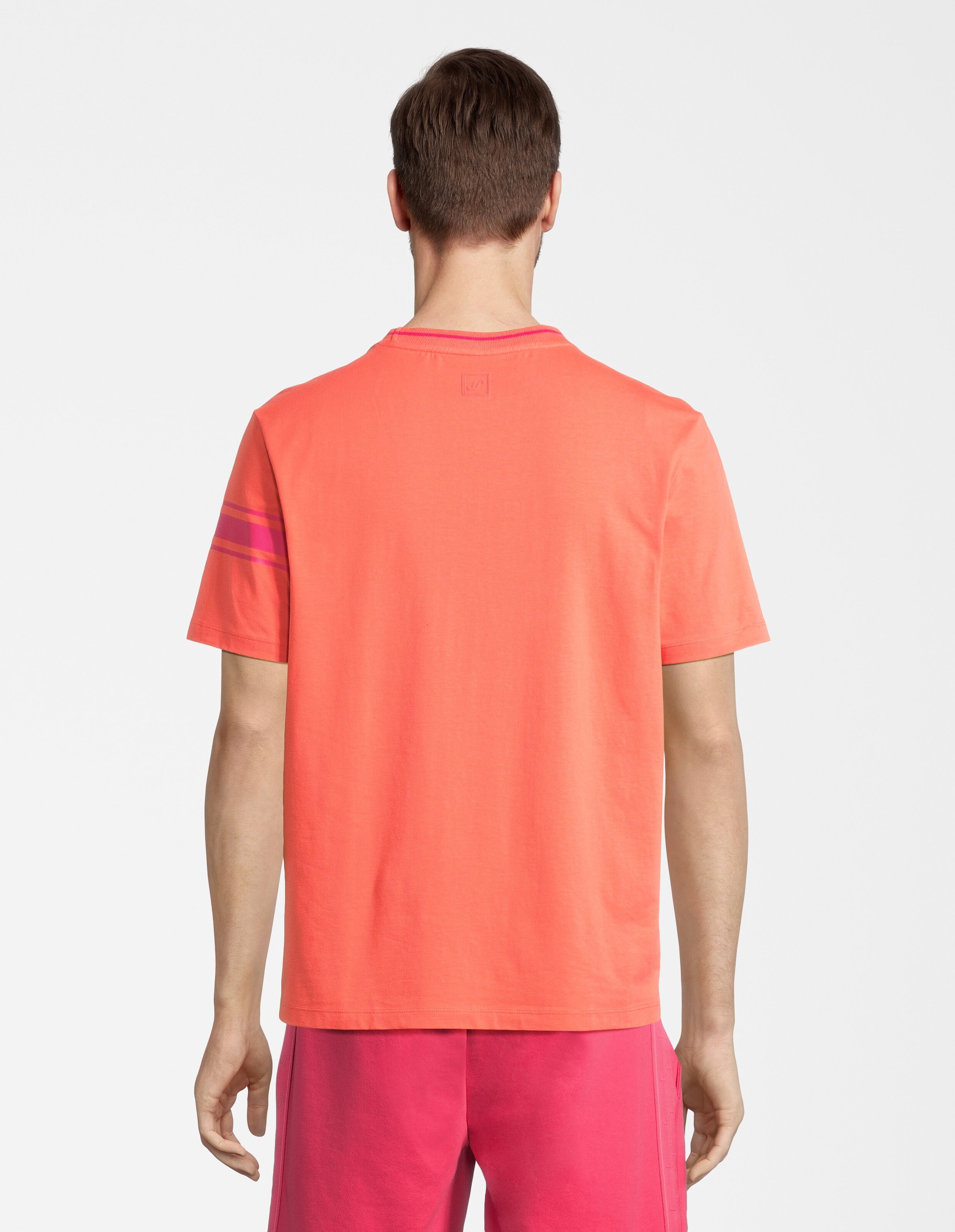 Rückansicht eines Mannes in leuchtend orangem T-Shirt mit Streifendesign am Ärmel und einer pinken kurzen Hose vor weißem Hintergrund.