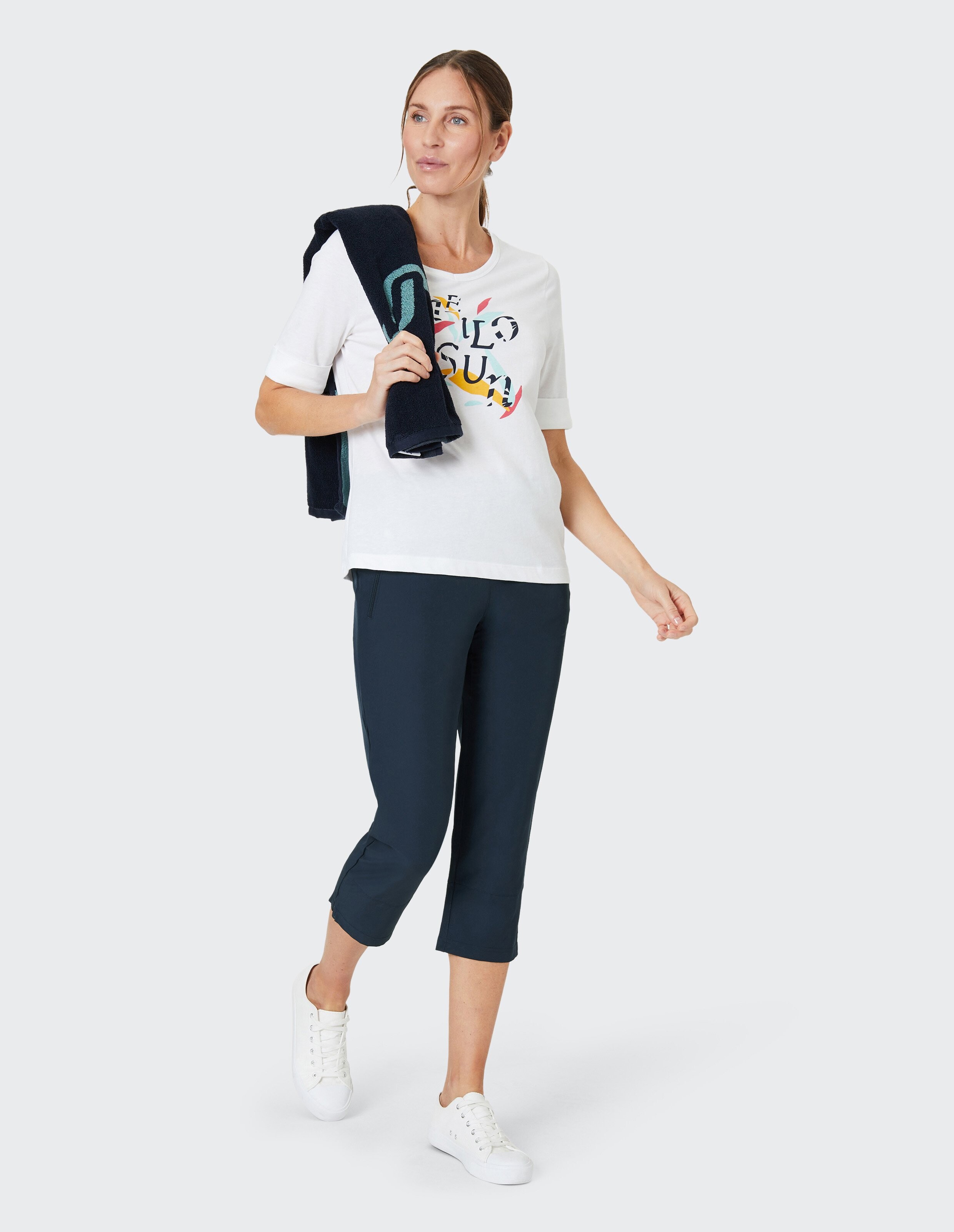 Frau mit weißem T-Shirt und buntem Schriftzug trägt dunkelblaue Jacke über der Schulter, kombiniert mit blauen Caprihosen und weißen Sneakers.