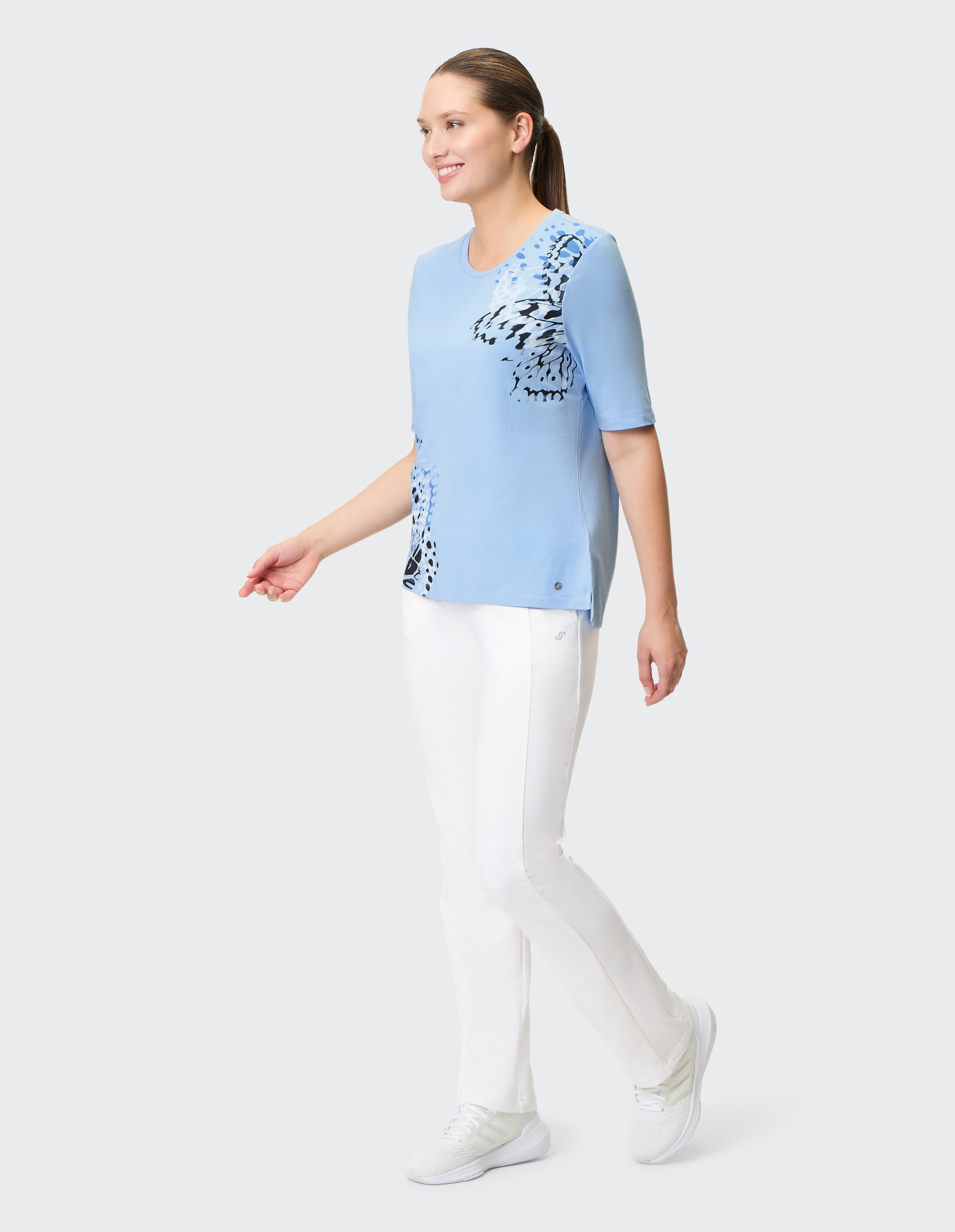 Frau trägt ein hellblaues T-Shirt mit Schmetterlingsdruck und eine weiße Freizeithose.