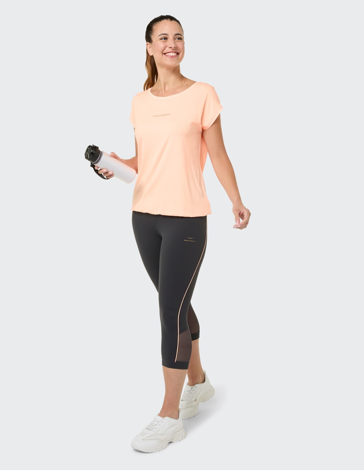 Frau in pfirsichfarbenem T-Shirt und schwarzen Leggings hält eine Wasserflasche in der Hand und lächelt.