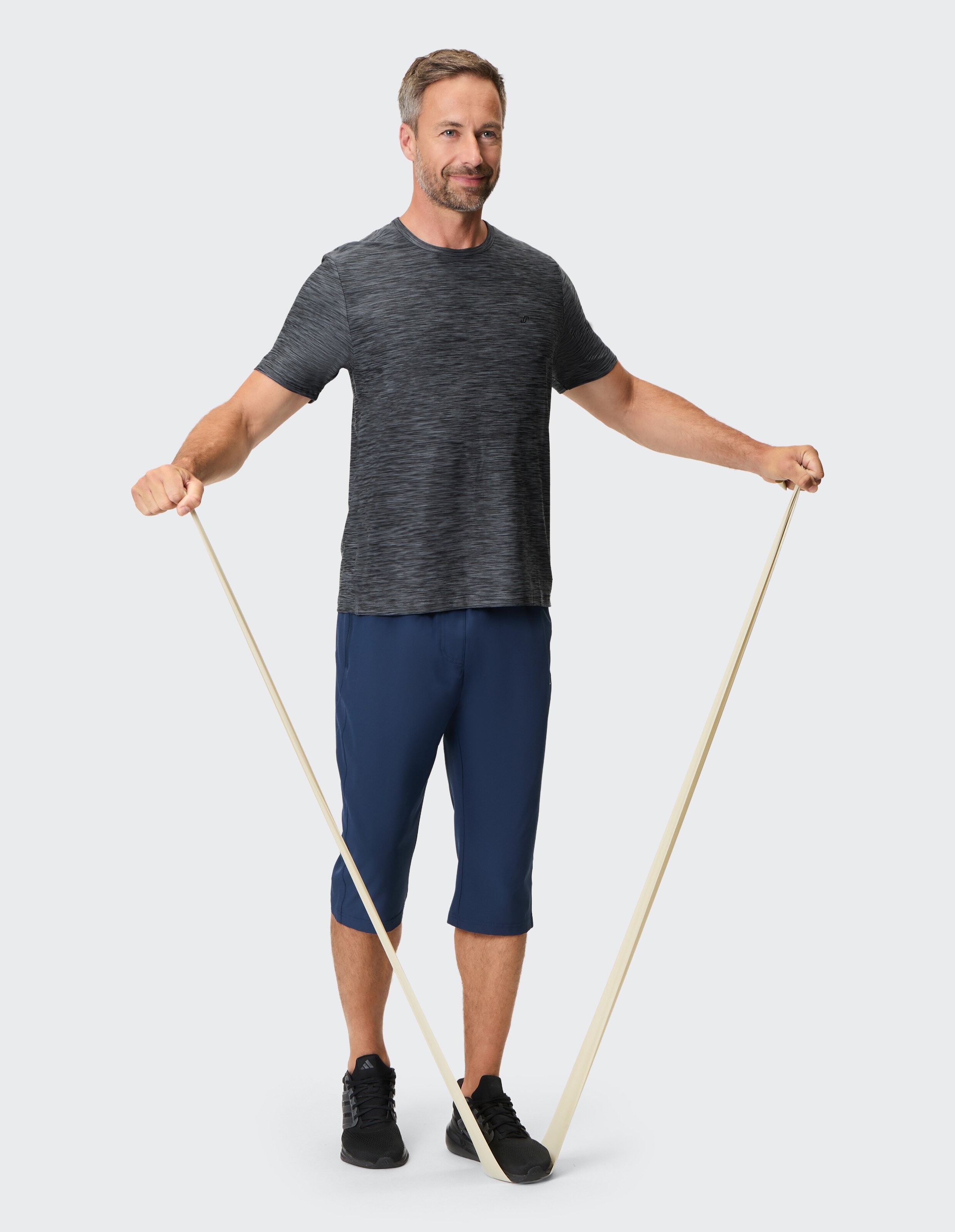 Ein Mann in Sportkleidung macht Übungen mit einem Widerstandsband; er steht aufrecht, hält das Band an beiden Enden gespannt.