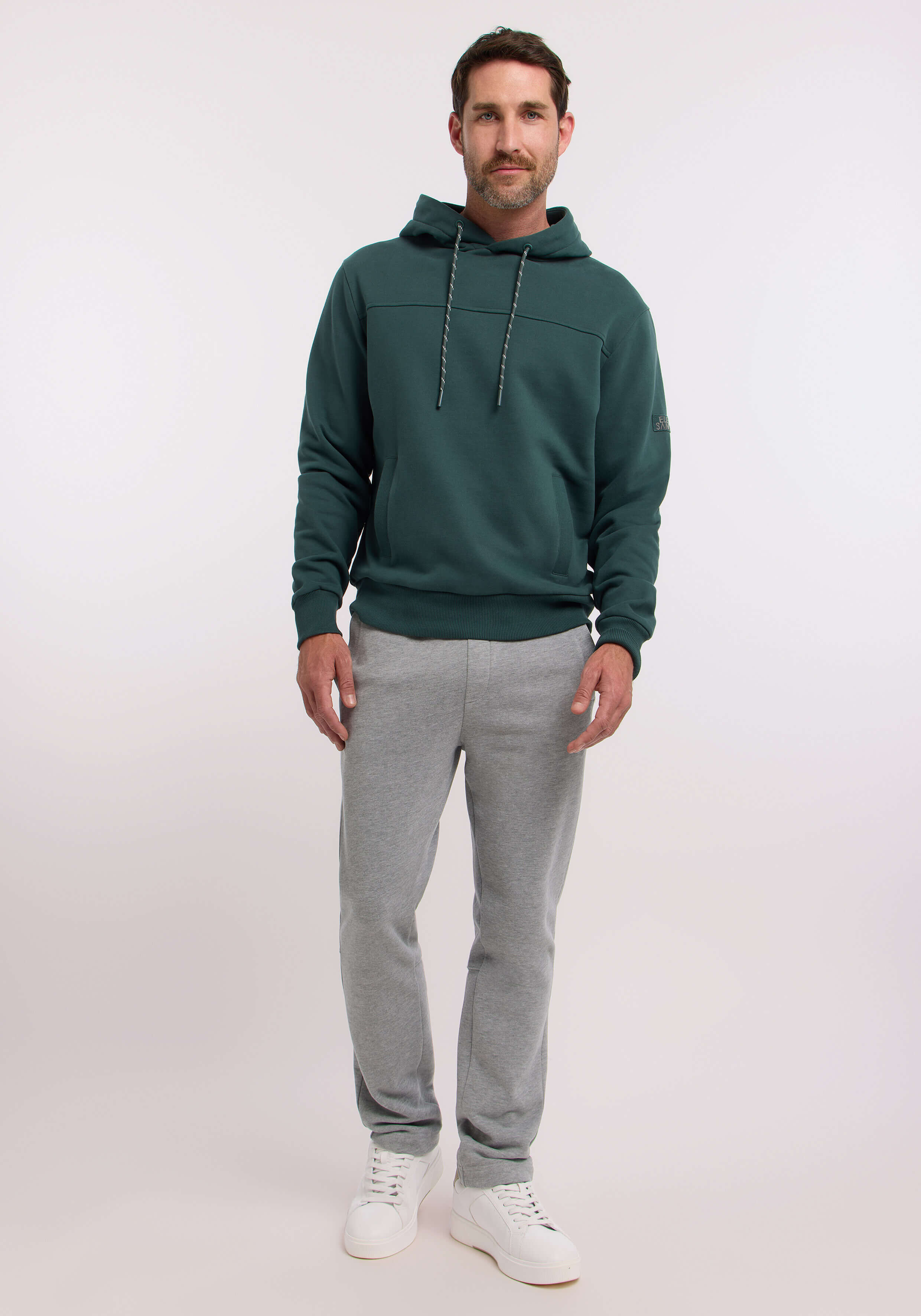 Mann trägt einen gemütlichen grünen Kapuzenpullover und eine graue Jogginghose, gestylt für lässigen Komfort.