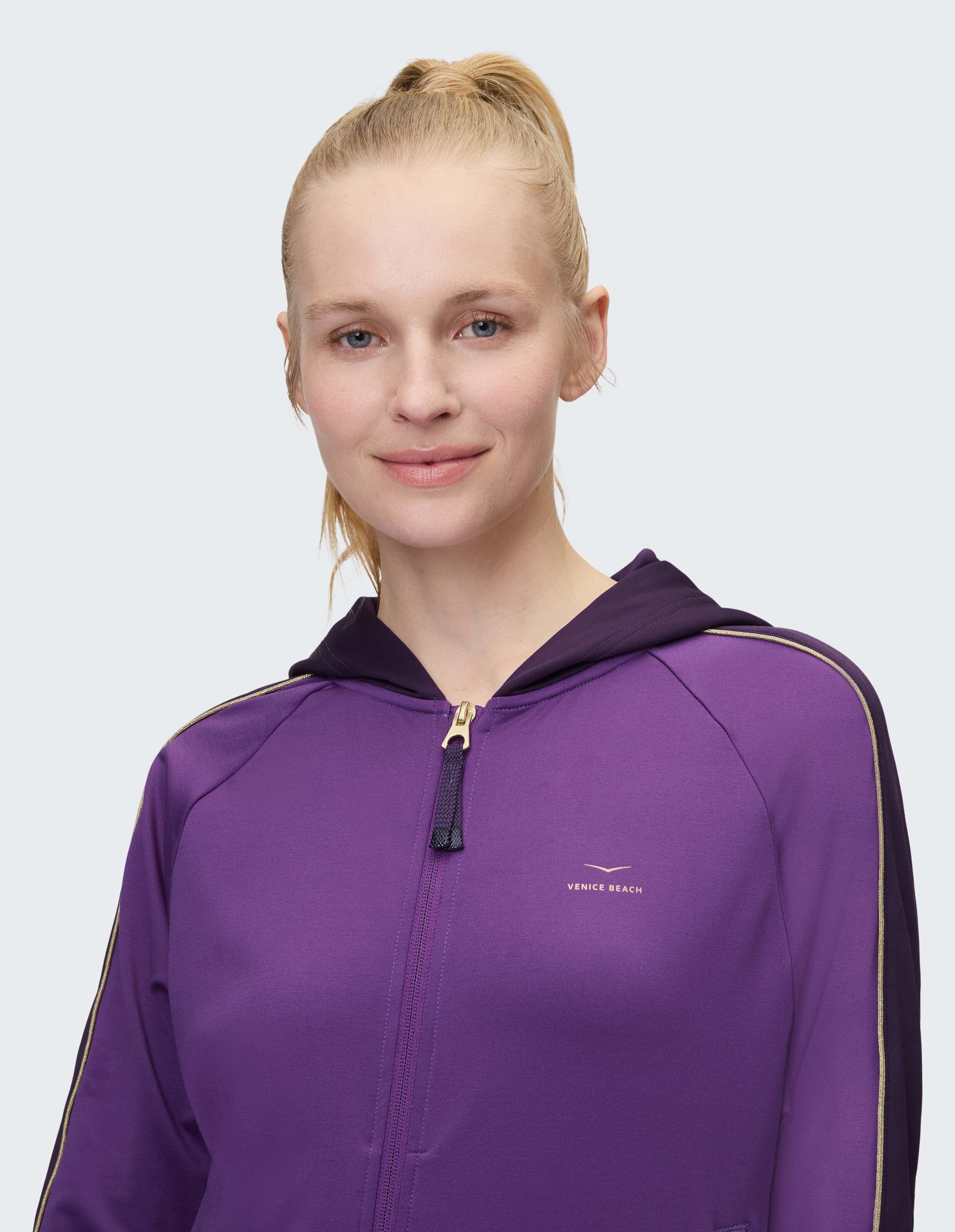 Frau in einem lilafarbenen Kapuzenpullover mit Reißverschluss und goldenen Akzenten.