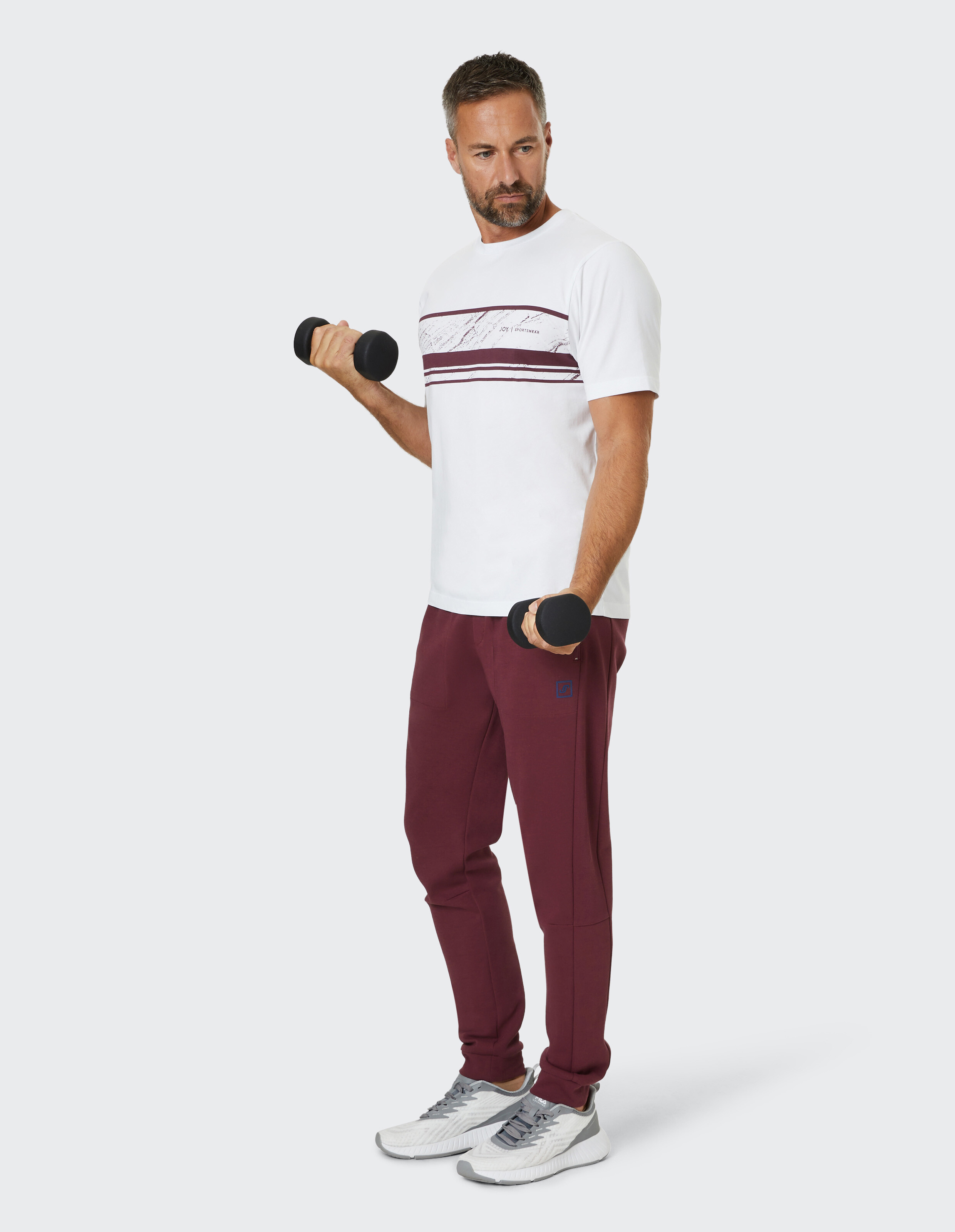 Mann in weißem T-Shirt und bordeauxroter Jogginghose hebt zwei Kurzhanteln, steht aufrecht, schaut zur Seite und trägt weiße Sportschuhe.
