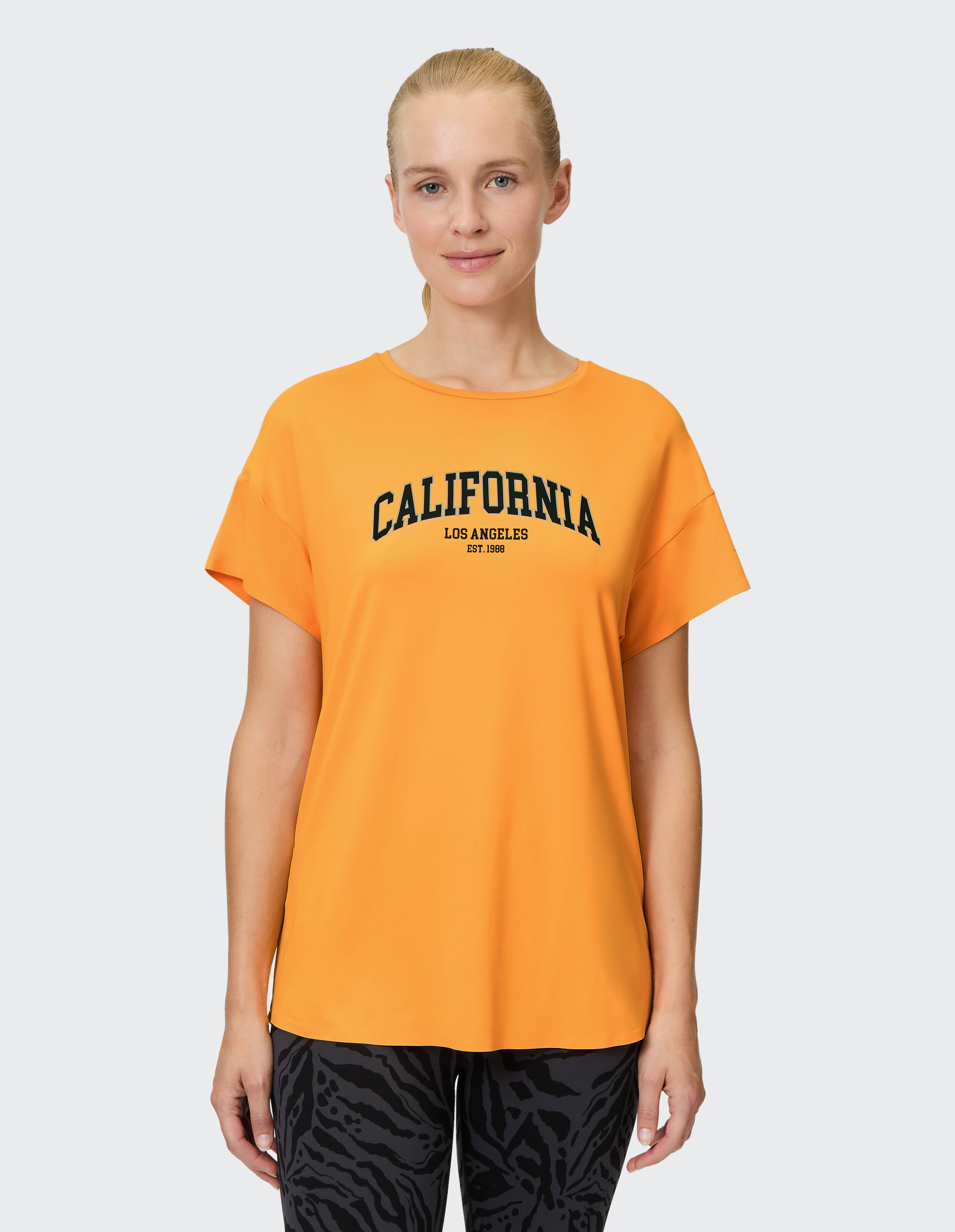 Frau trägt ein orangefarbenes Venice Beach Edana Kurzarmhemd mit California-Aufdruck.