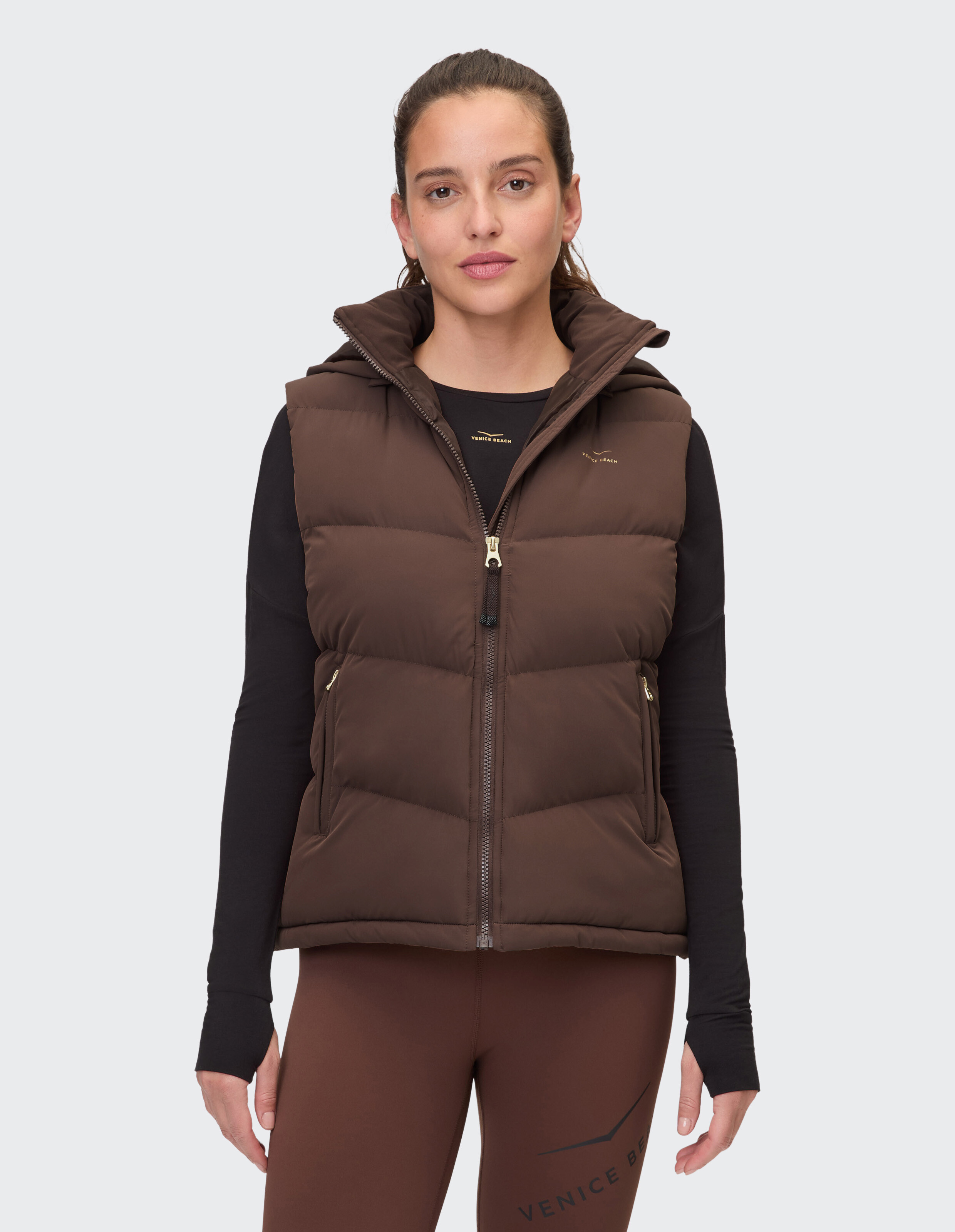 Braune Steppweste mit Kapuze, gepaart mit einem schwarzen Langarmshirt und braunen Leggings.