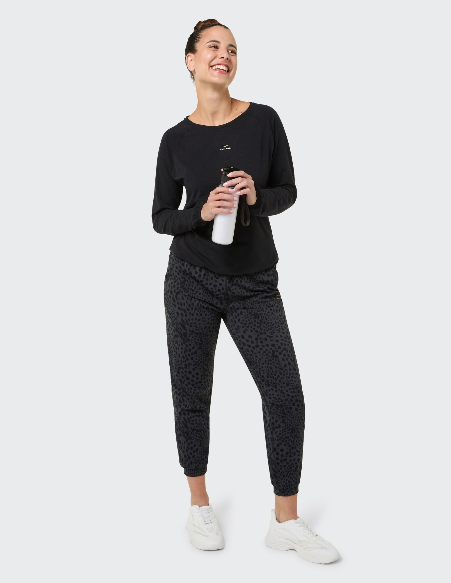 Lächelnde Frau in schwarzem Langarmshirt und Jogginghose mit Leopardenmuster, hält eine Wasserflasche in der Hand.