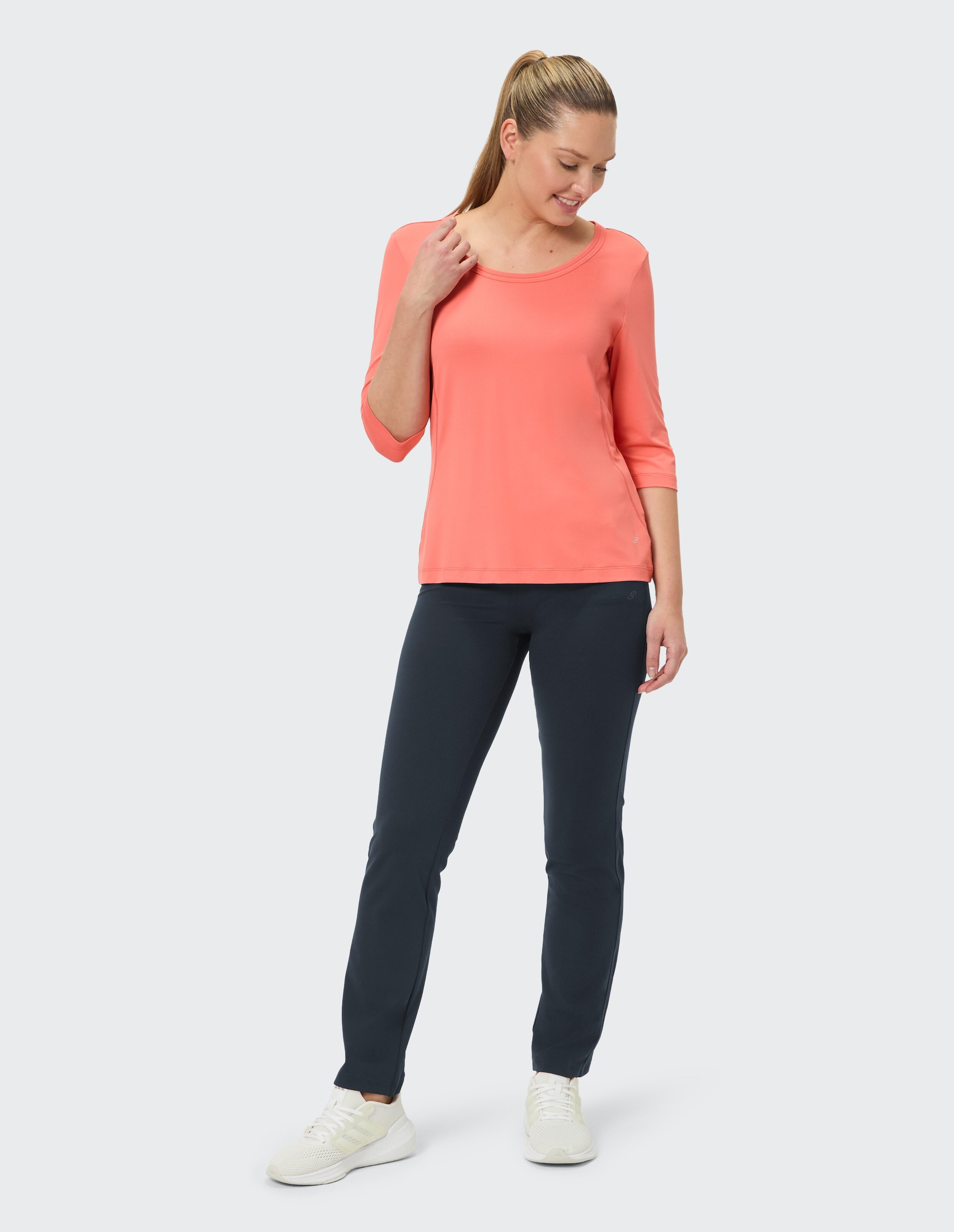 Frau in weißem T-Shirt mit Aufdruck und dunkler Hose steht mit Sneakers, schaut nach oben, Haare im Pferdeschwanz, entspannte Pose.
