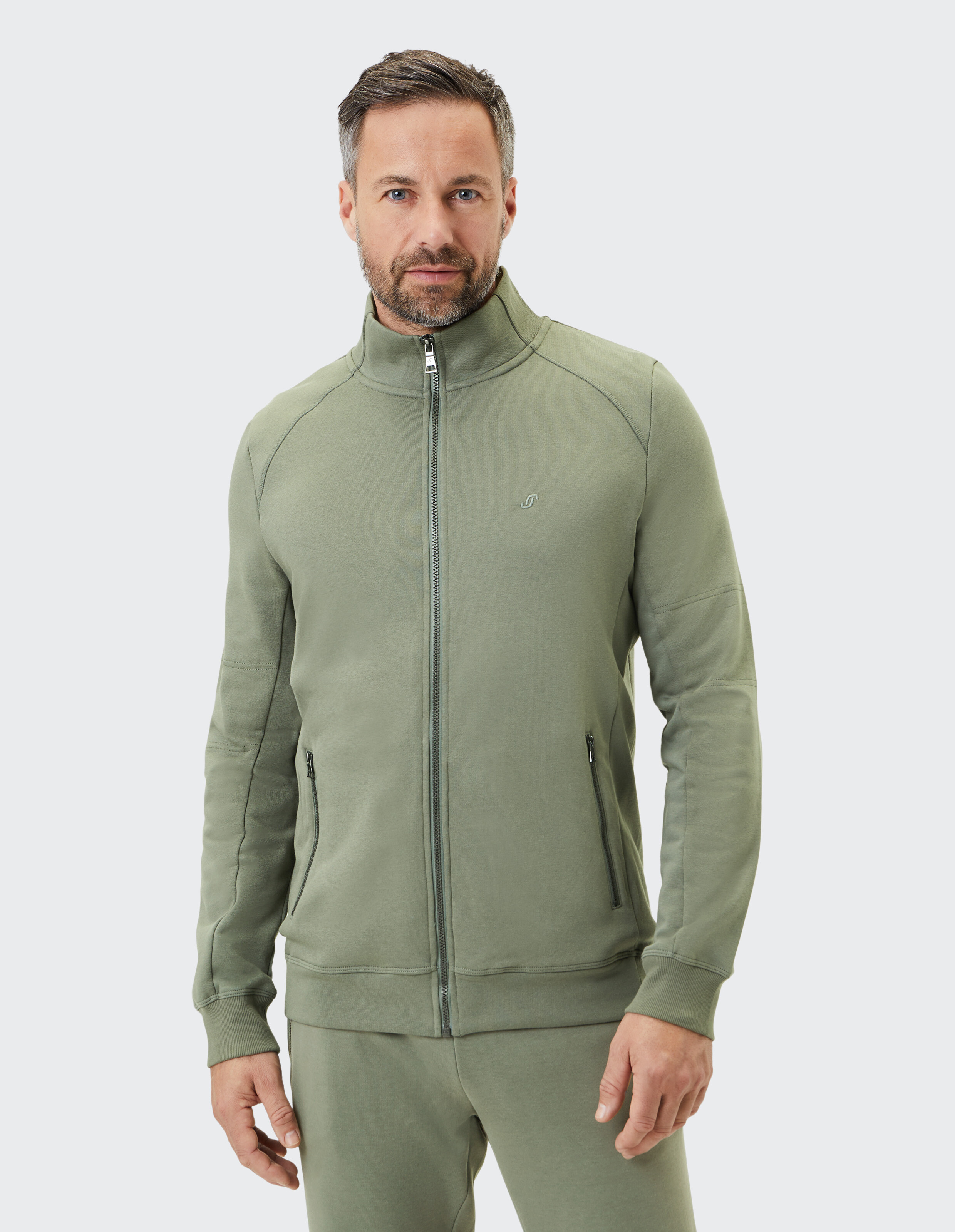 Ein Mann trägt einen olivgrünen Trainingsanzug mit einem Reißverschluss und zwei Taschen, vor neutralem Hintergrund.