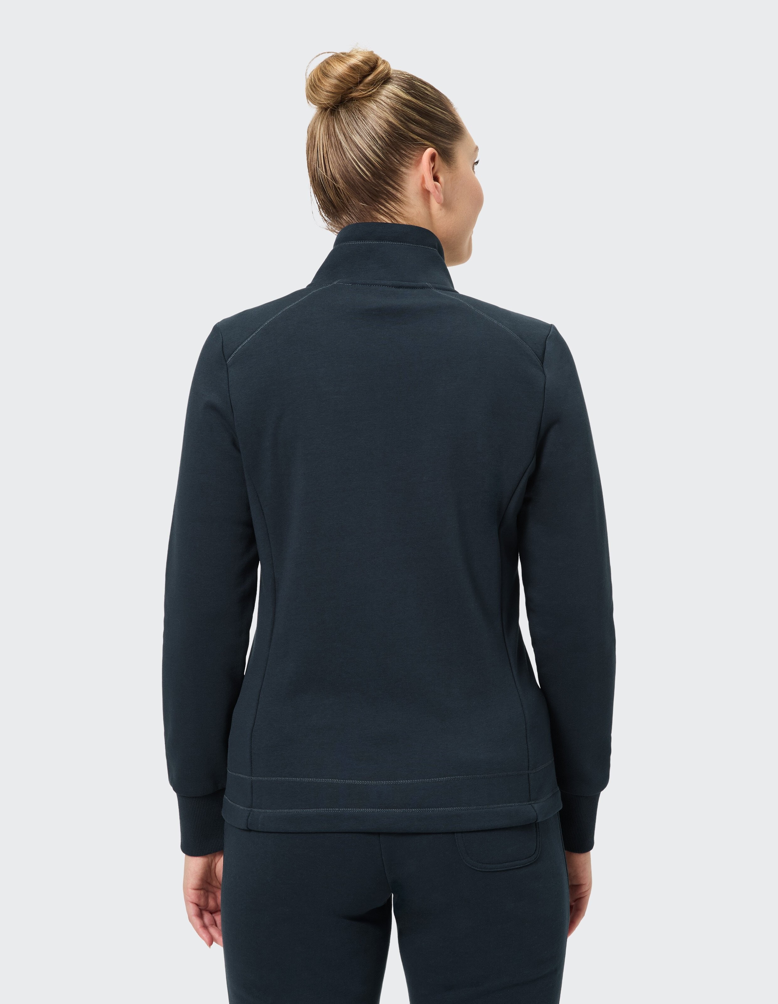 Eine Frau mit langen braunen Haaren im Pferdeschwanz trägt eine dunkelblaue Jacke und Hose, stehend mit dem Rücken zur Kamera.