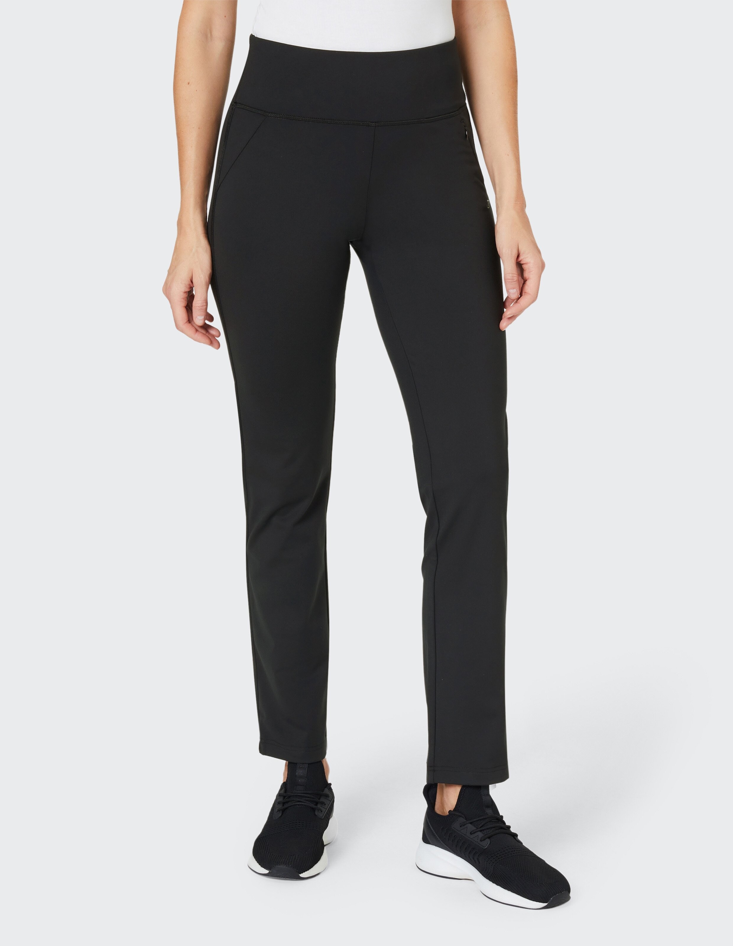 Eine Person trägt eine schwarze, taillierte Sporthose mit hohem Bund und Taschen, kombiniert mit schwarzen Sneakers auf grauem Hintergrund.